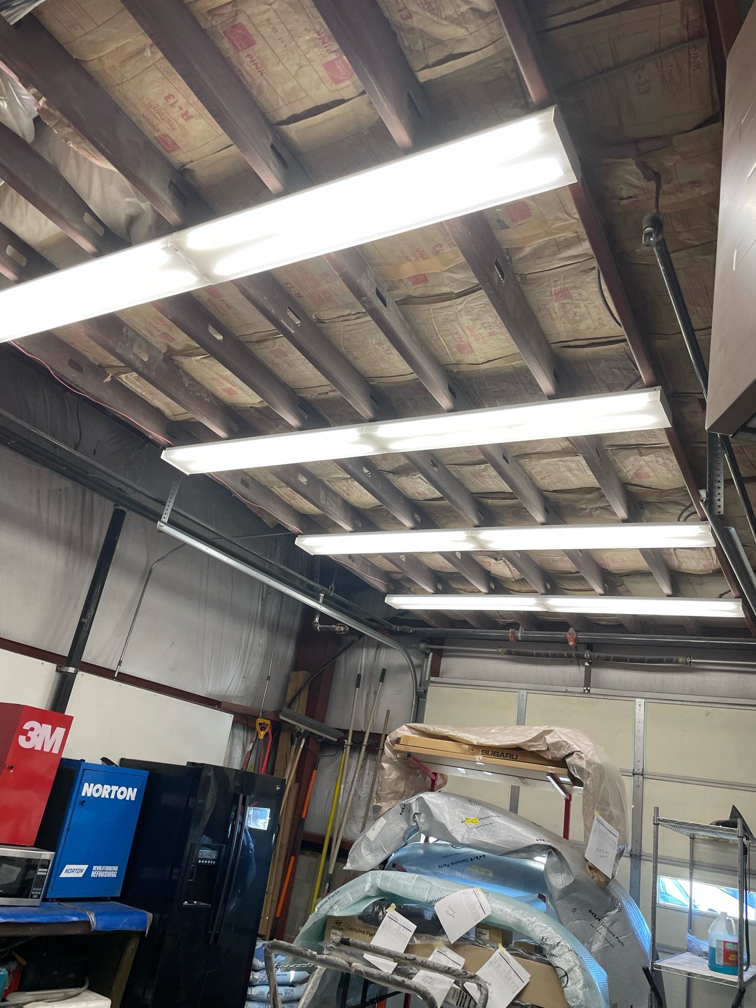 Interior of a garage or workshop showing fluorescent lights, insulation, a red 3M box, a blue Norton machine, and various tools and covered items.