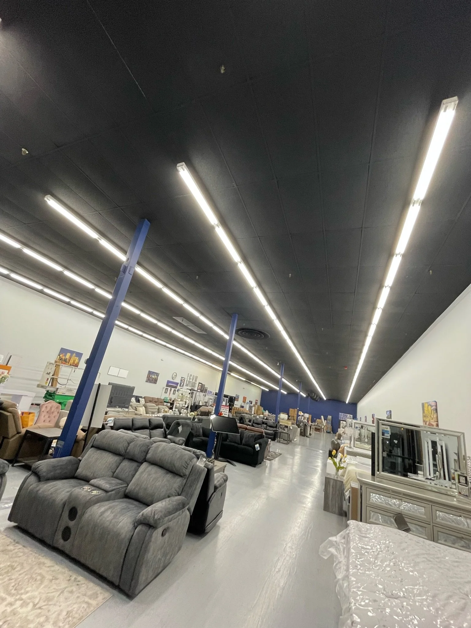 Interior of a furniture store with various sofas, chairs, and tables on display, illuminated by long ceiling lights, with a dark ceiling and light-colored floor.