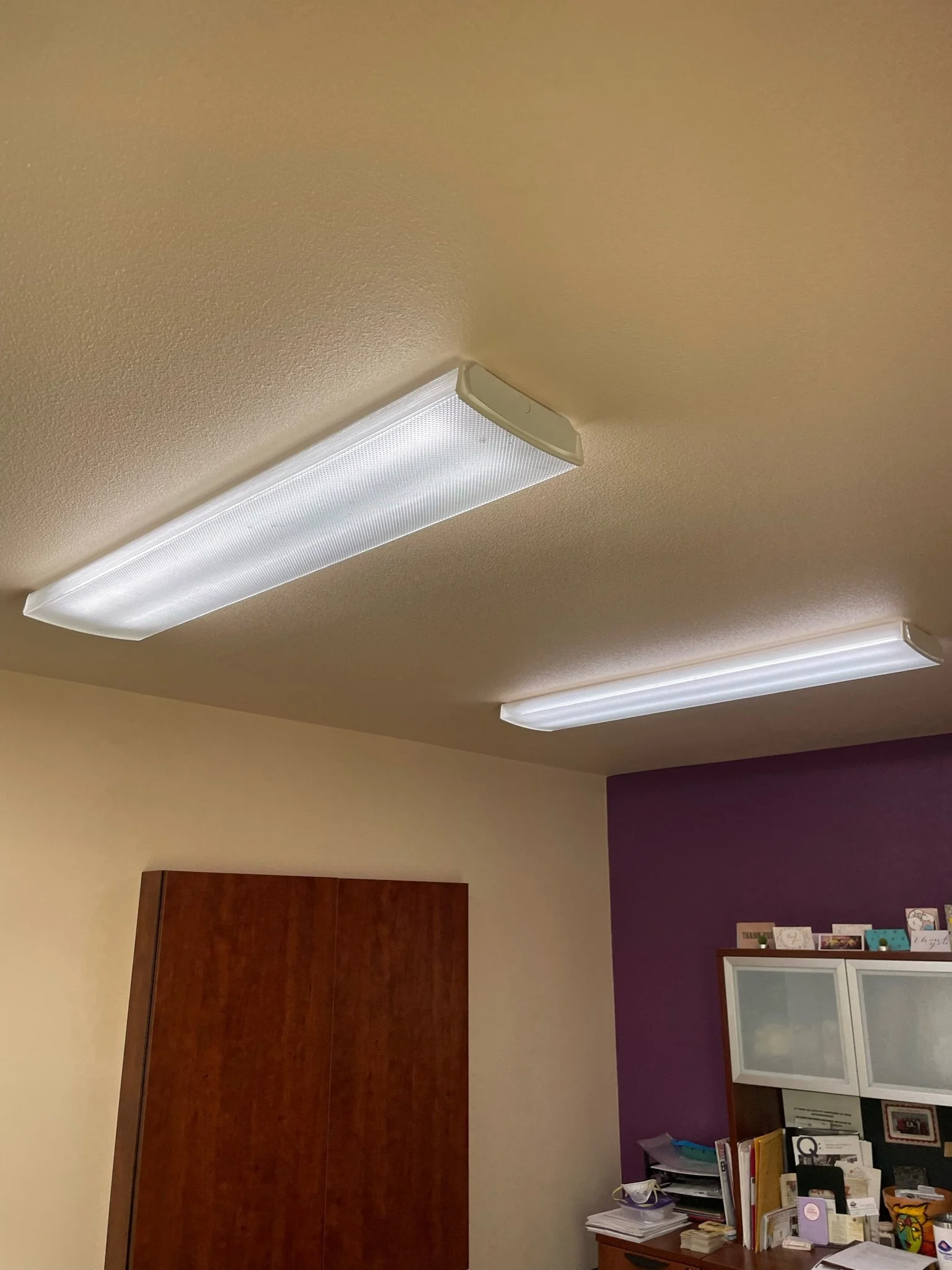 Ceiling with two fluorescent light fixtures in a room with beige and purple walls, a wooden cabinet, and a desk with papers and decorations.
