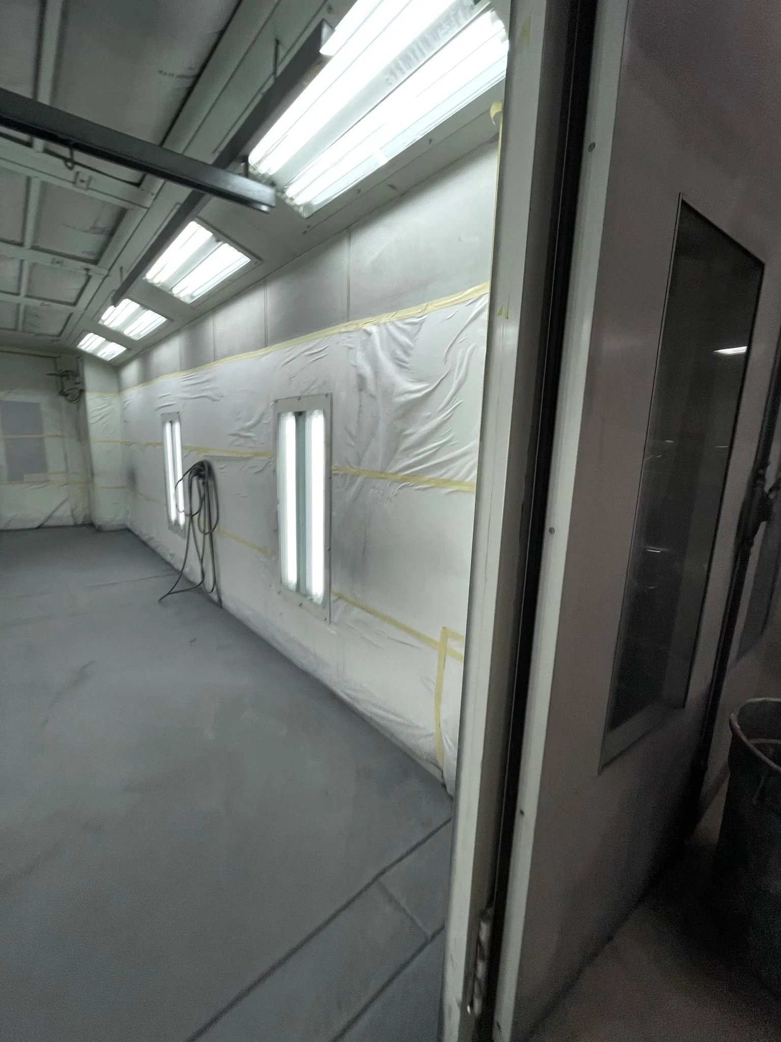 An interior view of a spray booth with plastic sheeting and bright fluorescent lights, including walls, ceiling, and a doorway.