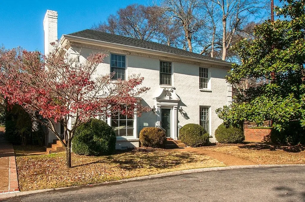 A two-story white house with multiple windows, surrounded by trees and shrubs, with a small set of brick steps leading to the front door, set in a residential neighborhood under a blue sky.