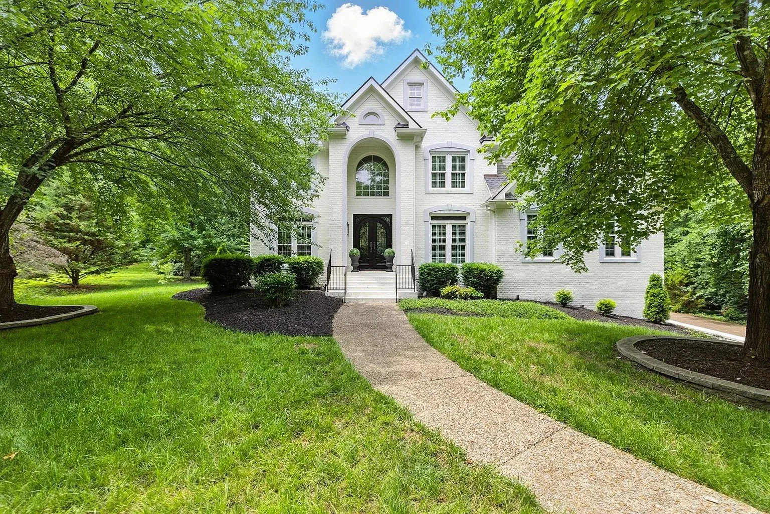 A white, two-story house with large front windows and a black front door. The house is surrounded by a well-maintained green lawn with trees and bushes, and a concrete pathway leading up to the front stairs.
