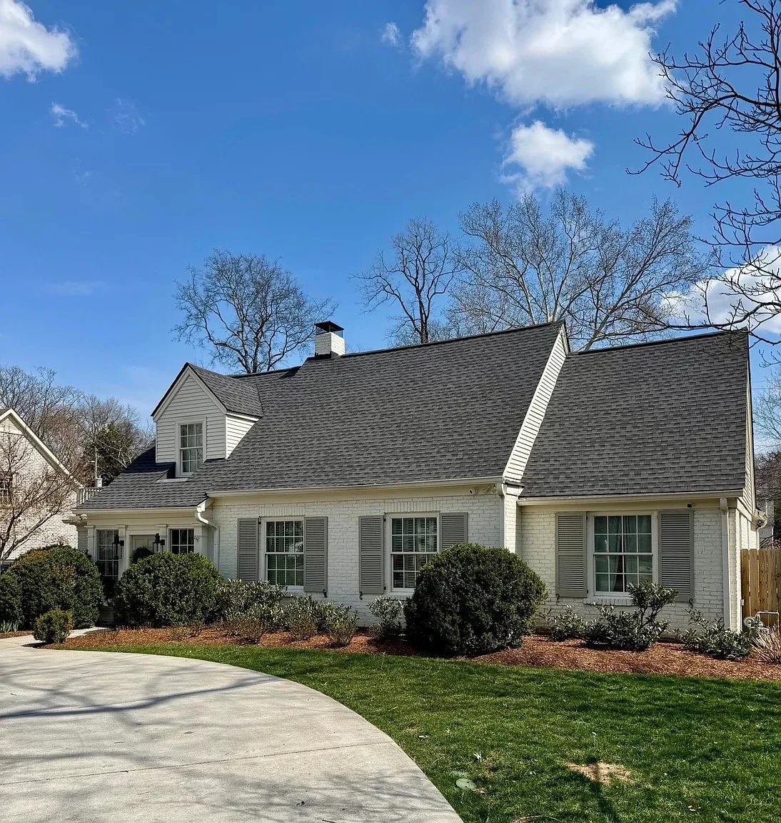 A white house with a dark gray shingle roof, multiple windows with shutters, surrounded by bushes, under a blue sky with some clouds.