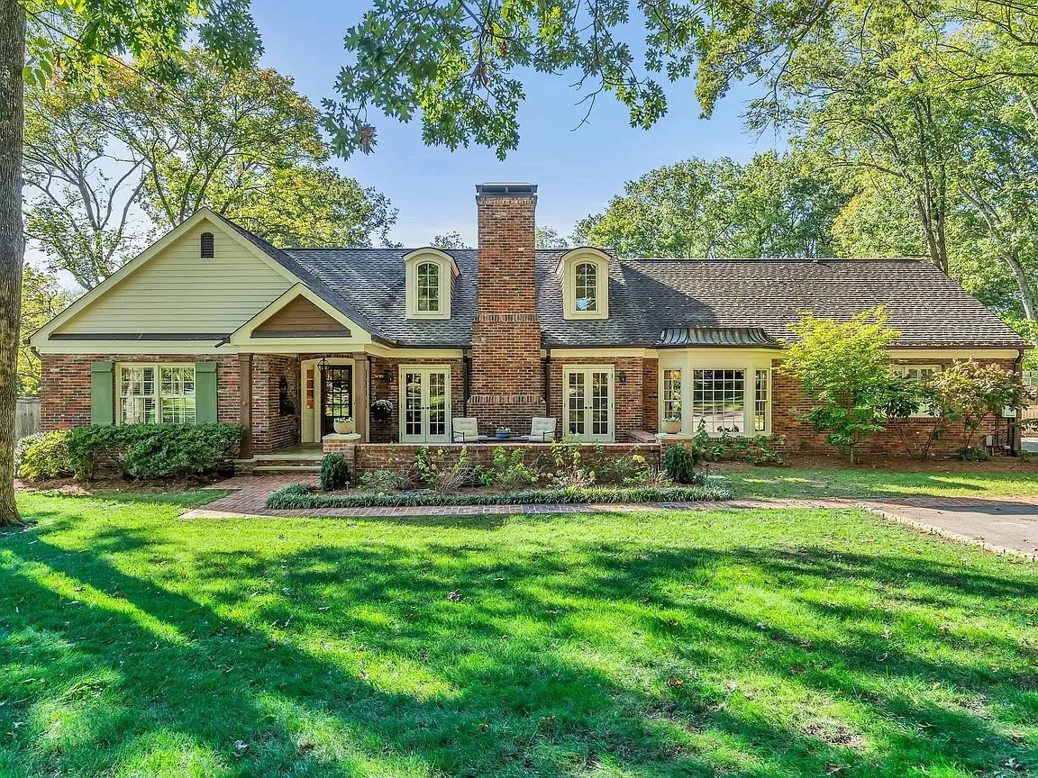 A house with a brick exterior, green shutters, and a tiled roof, surrounded by a well-maintained lawn and trees.