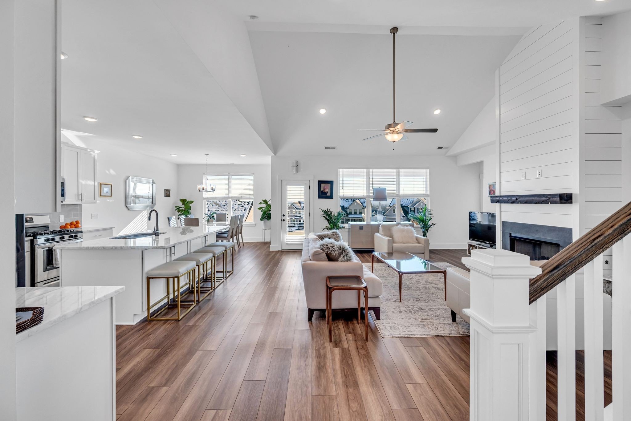 Open-concept living room and kitchen with hardwood flooring, white walls, and modern furniture. The living room has a white sofa, a cream armchair, a wooden coffee table, and a TV near a fireplace. The kitchen features a large white island with barstools, white cabinetry, and stainless steel appliances. Large windows let in natural light.