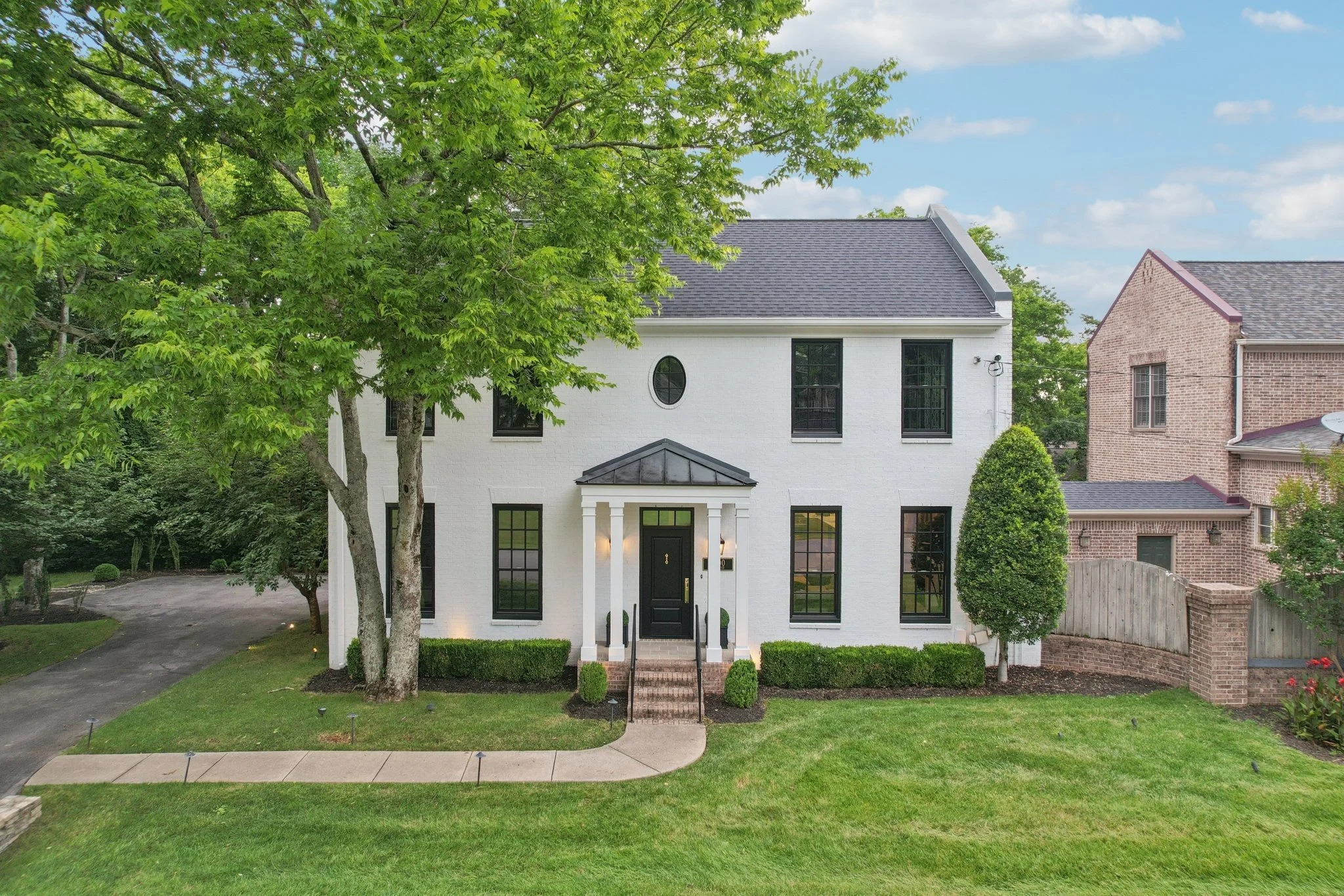 A white two-story house with black windows, a black front door, and a small front porch with brick steps. The house is surrounded by green grass, a large tree on the left, and a smaller tree on the right. A curved sidewalk leads to the front steps, and neighboring houses are visible on the right.