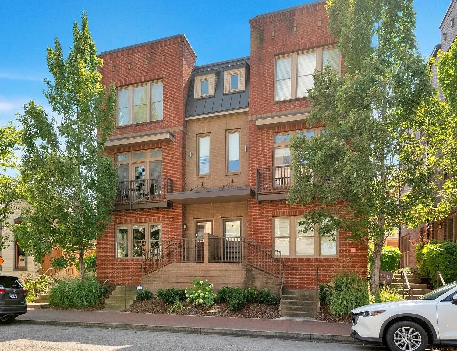 A multi-story residential building with brick and beige siding, featuring several windows and small balconies. There are trees and shrubs in the front yard, and two cars parked in front on the street.