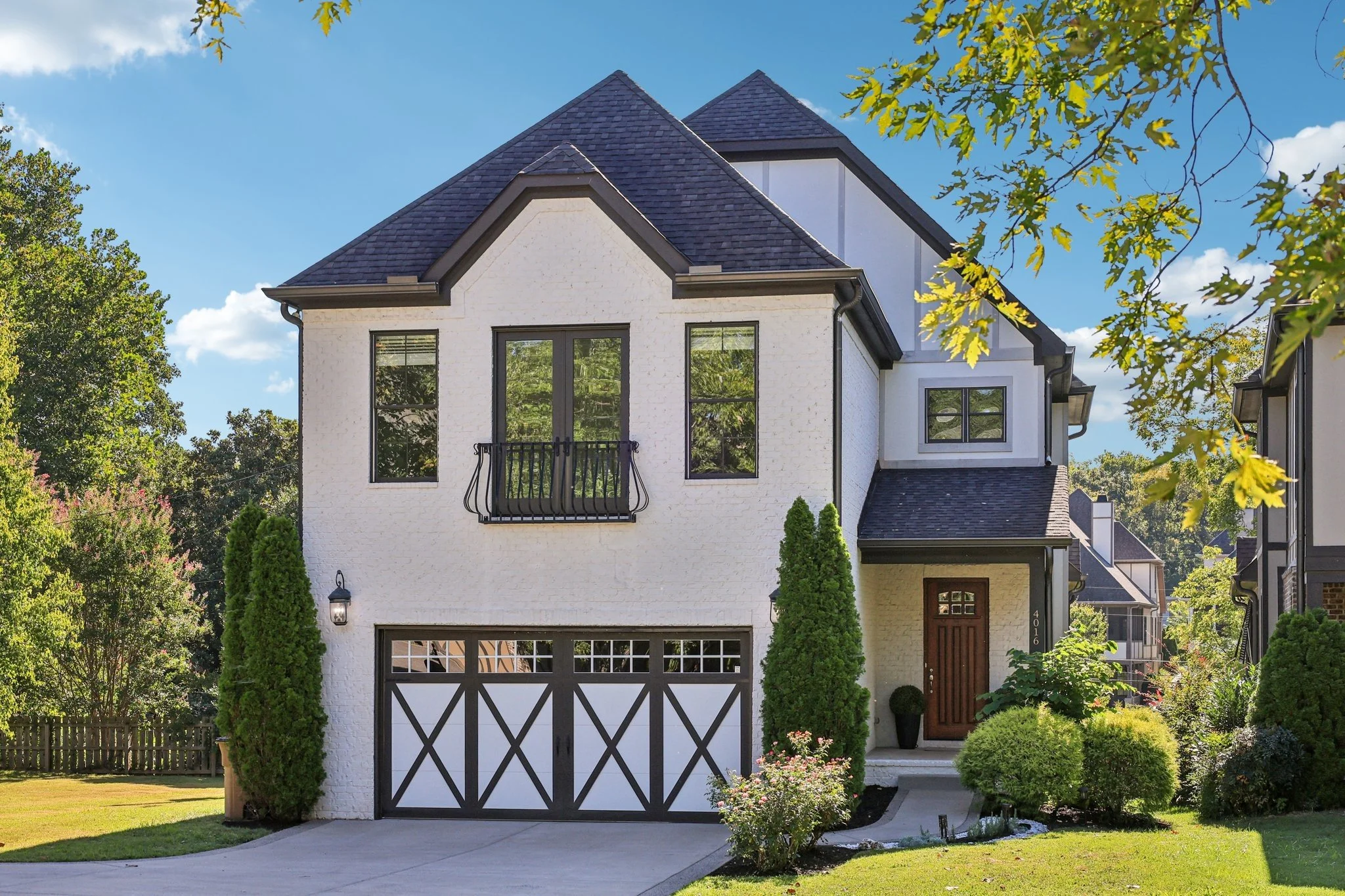 White two-story house with black roof, black window frames, and a black garage door with white trim, surrounded by a lush green lawn and landscaped bushes under a blue sky.
