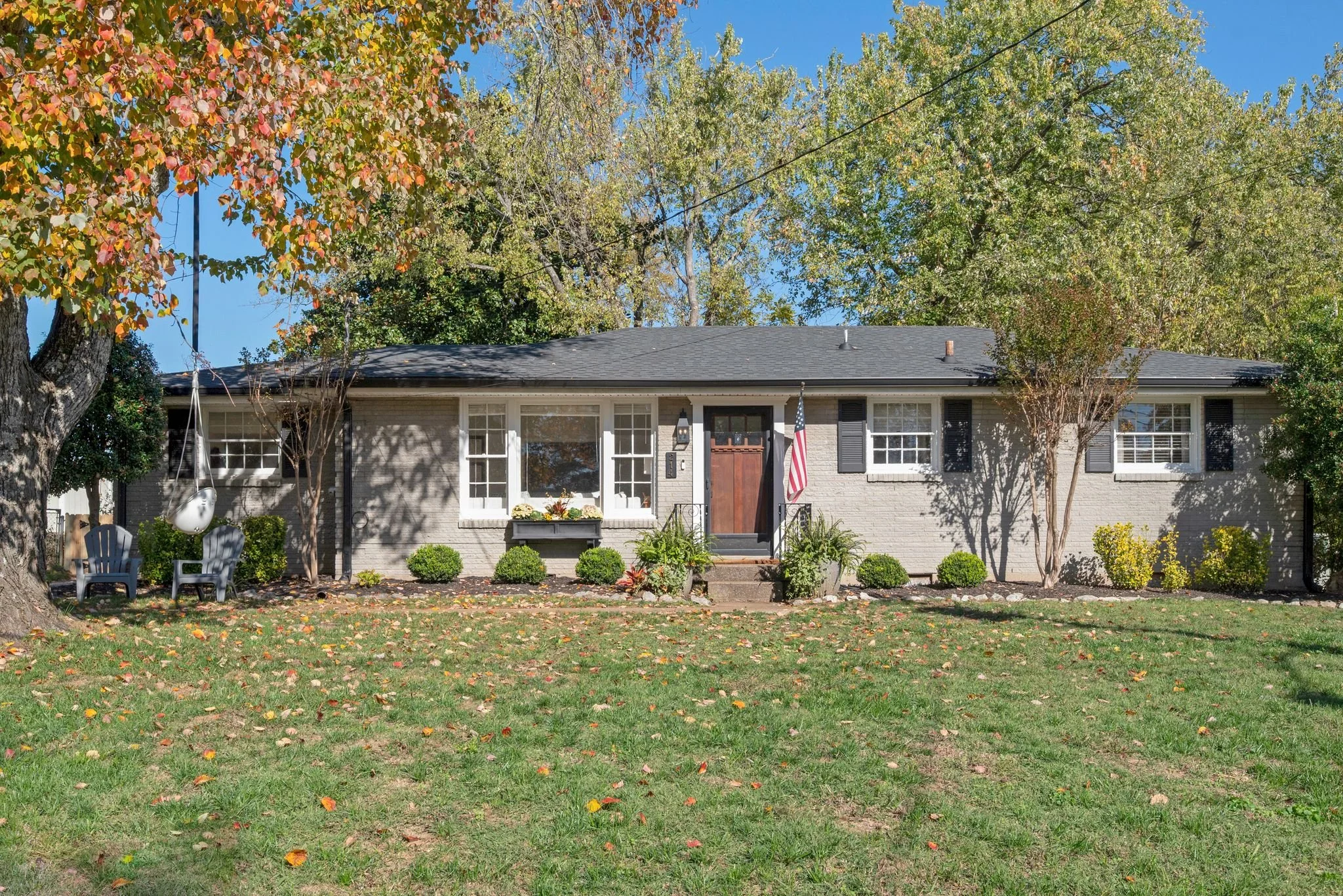 A white two-story house with black windows, a black front door, and a small front porch with brick steps. The house is surrounded by green grass, a large tree on the left, and a smaller tree on the right. A curved sidewalk leads to the front steps, and neighboring houses are visible on the right.