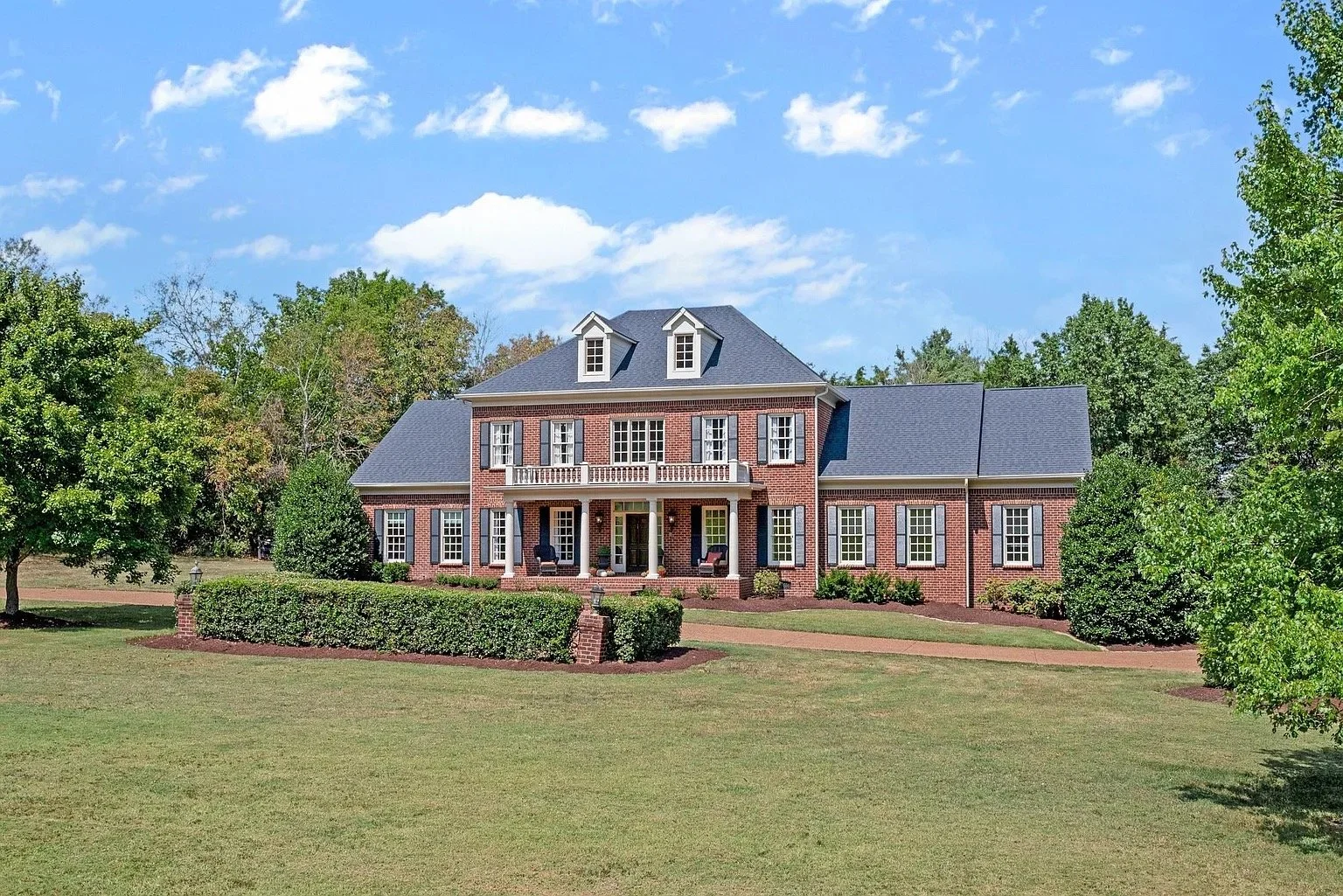 Large brick house with multiple windows, a front porch with columns, and a balcony, surrounded by green trees and a lawn under a blue sky with clouds.