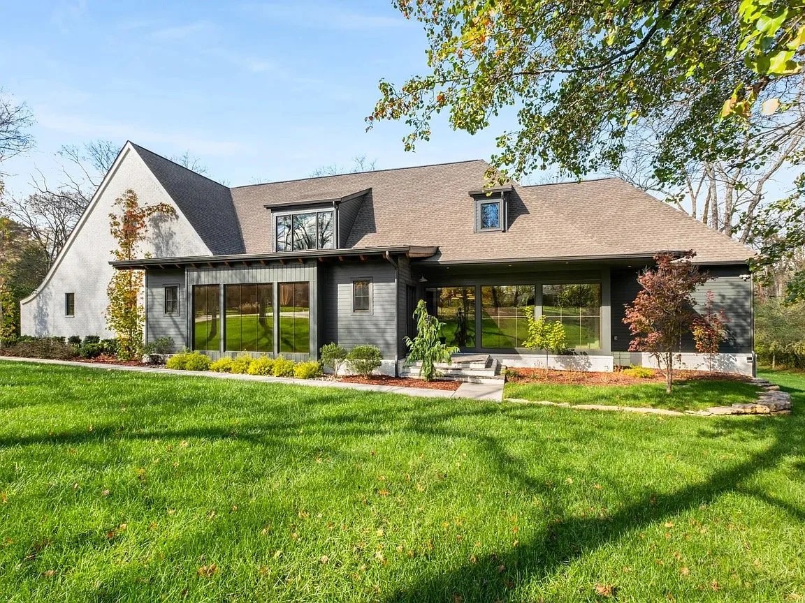 A modern two-story house with black siding and large windows, green lawn, and trees in the yard under a clear blue sky.