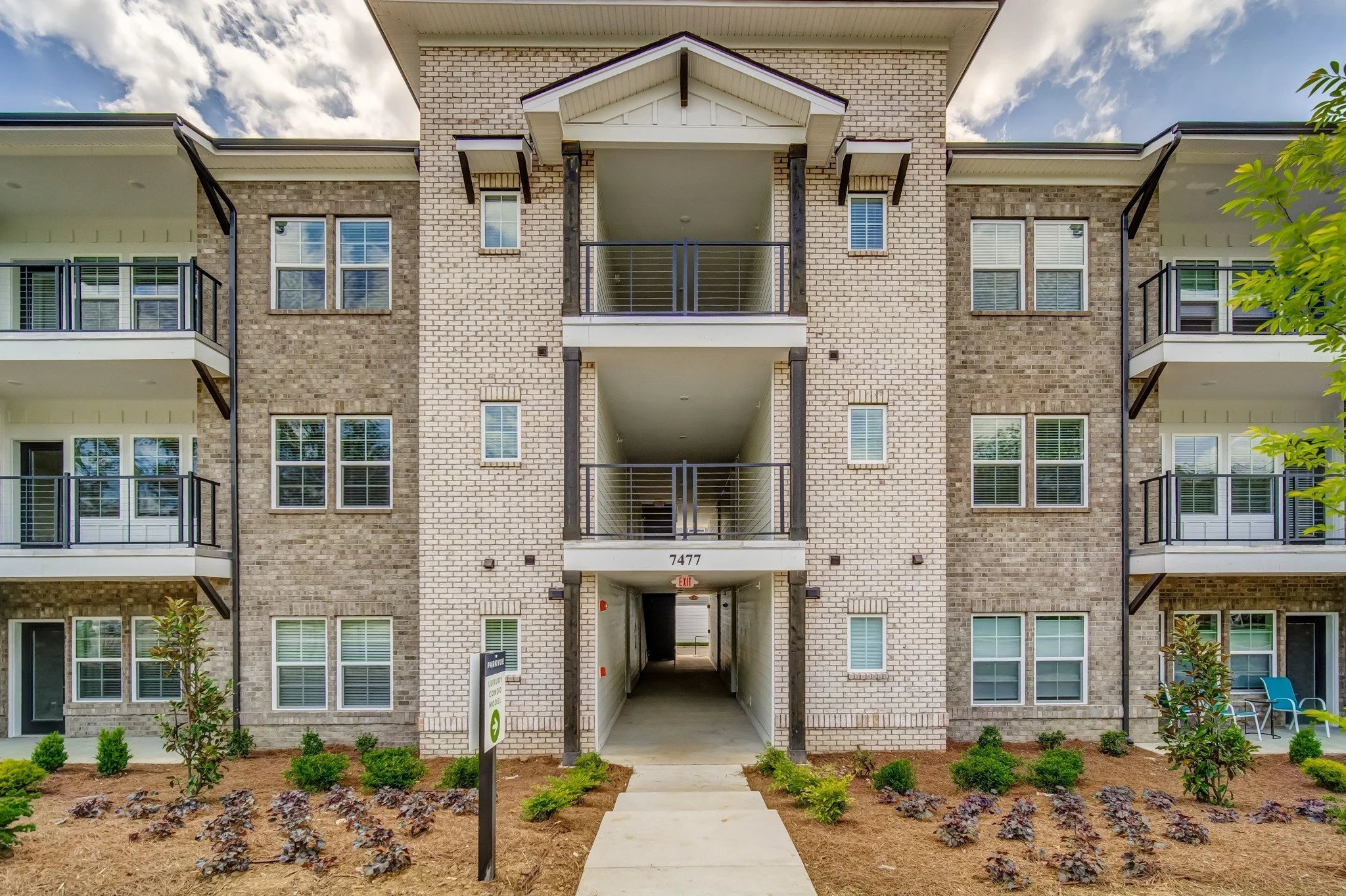 Exterior view of a modern, multi-story residential apartment building with balconies, brick and siding facade, and a landscaped front yard with small bushes and plants.