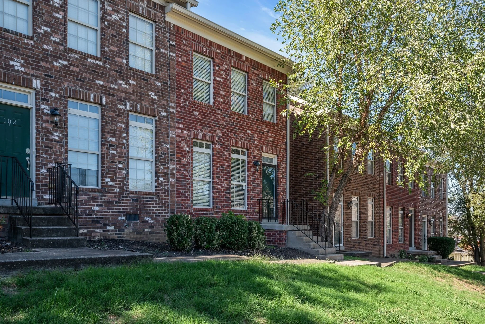 View of a row of brick apartments with staircases leading to front entrances, surrounded by green lawn and trees under a clear blue sky.