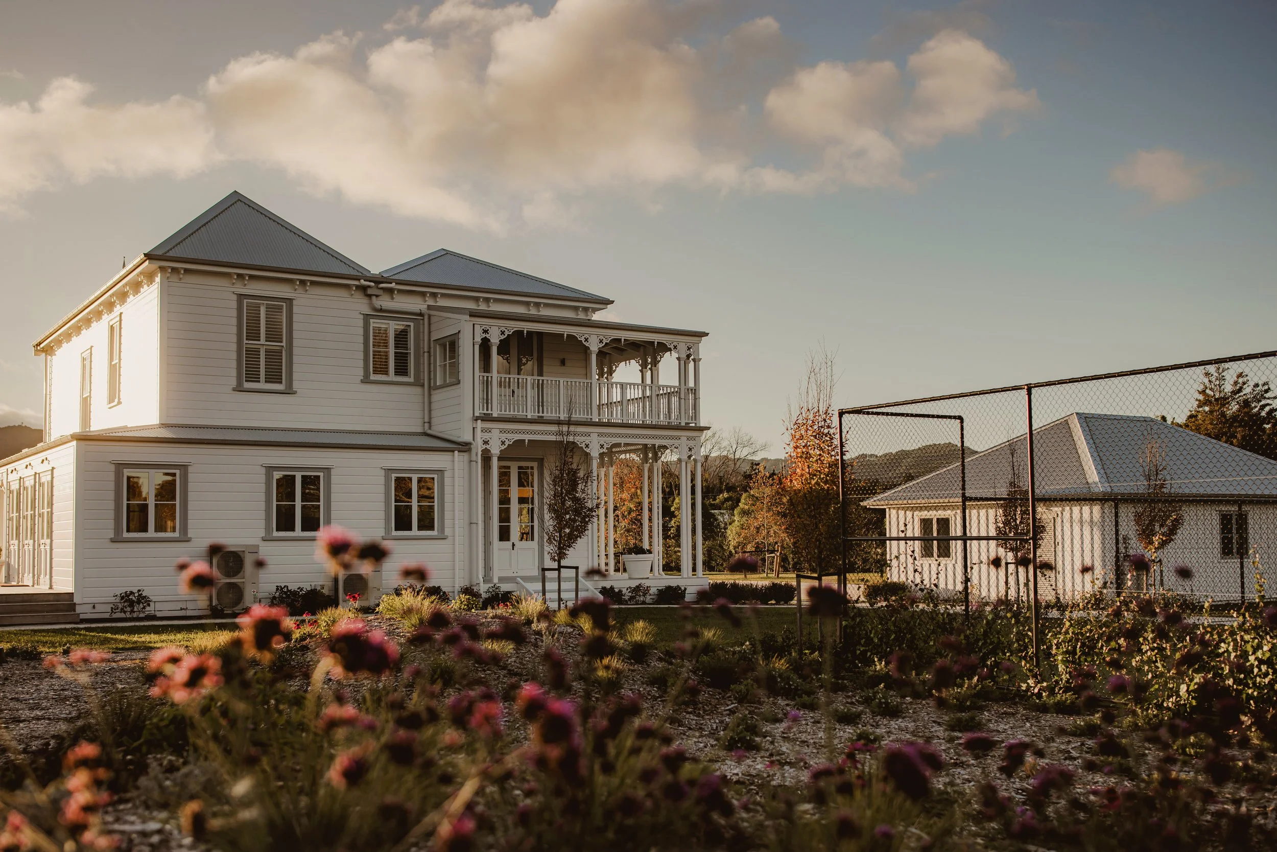 Victorian-style two-story house with ornate balcony, white exterior, surrounded by garden with blooming flowers and adjacent smaller building, under a blue sky with clouds.