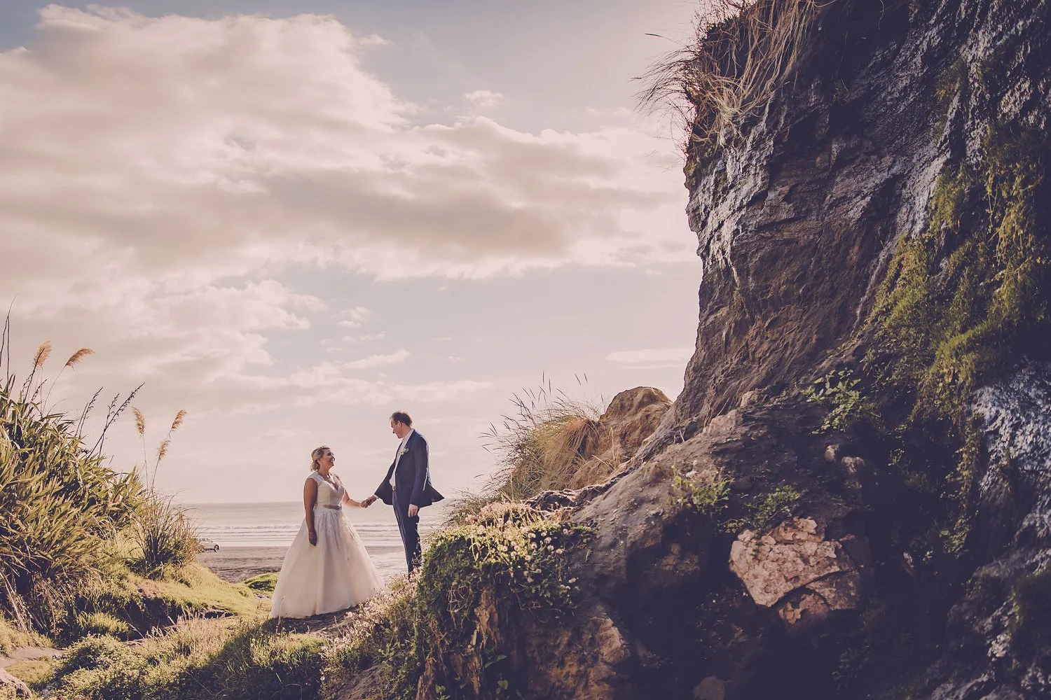 Bride and groom holding hands by coastal rock formation, ocean in background