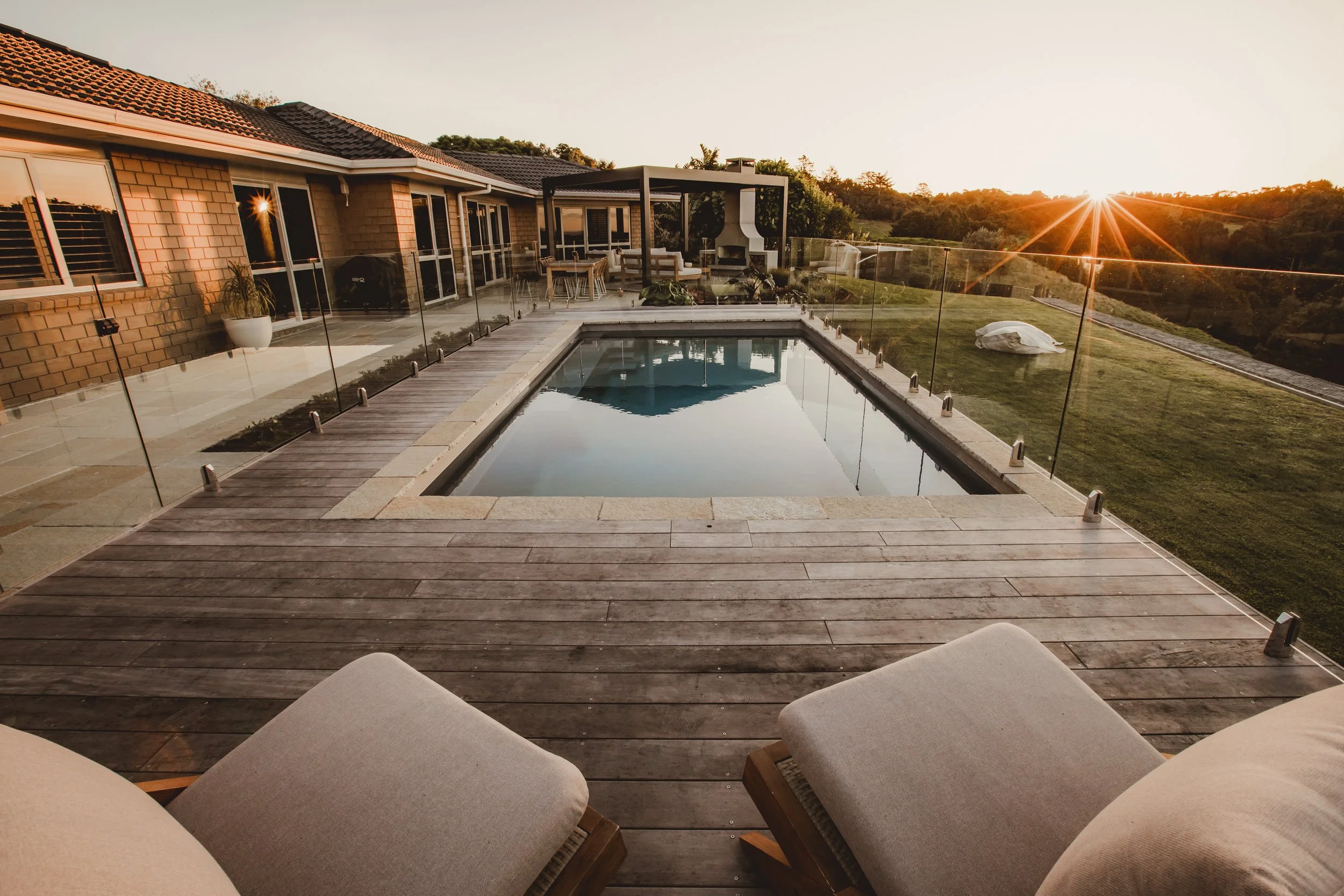 Backyard with swimming pool, wooden deck, and lounge chairs; house exterior; sunset view.