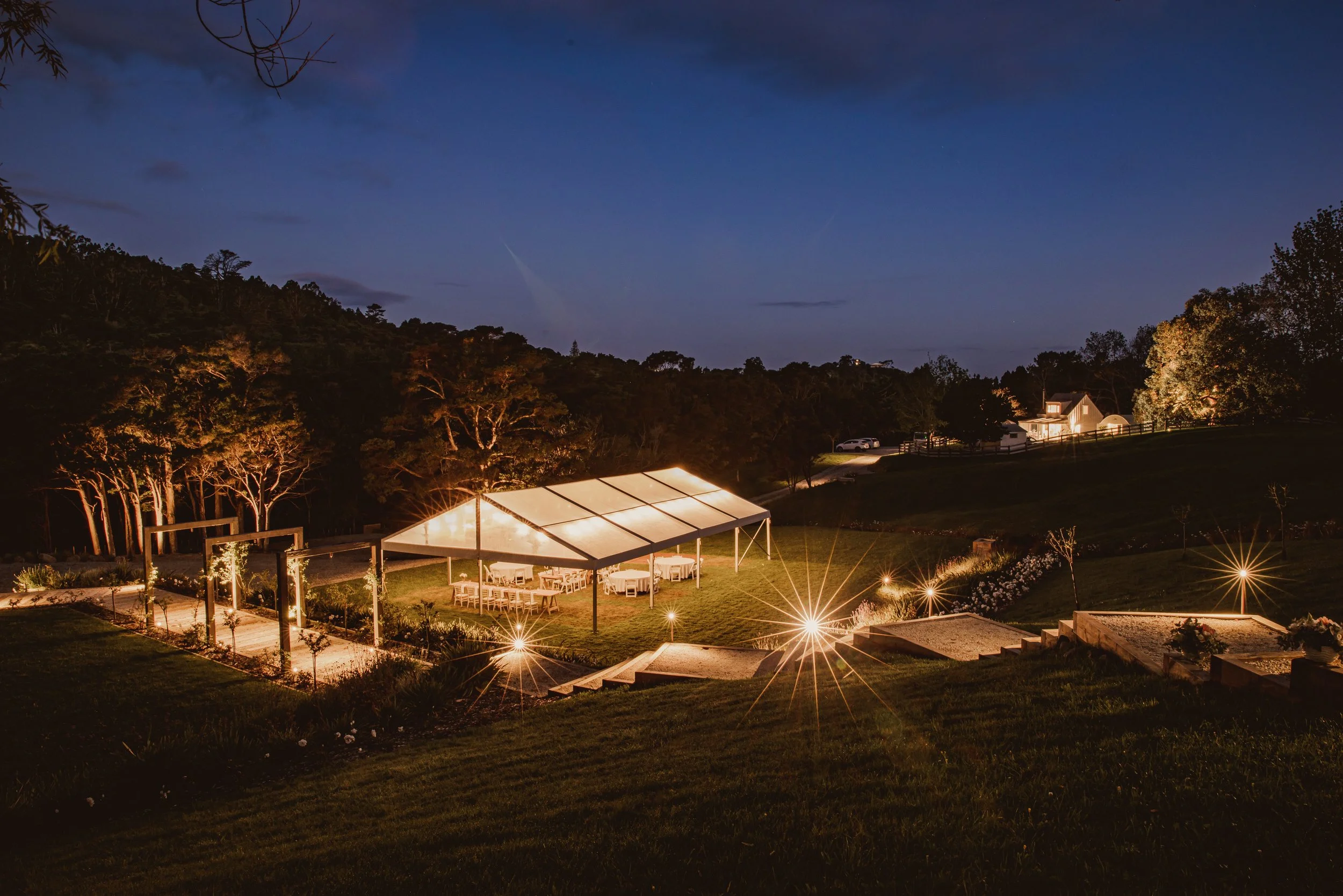 Outdoor event venue at night with a lit canopy, tables, chairs, surrounding trees, and a house in the distance.