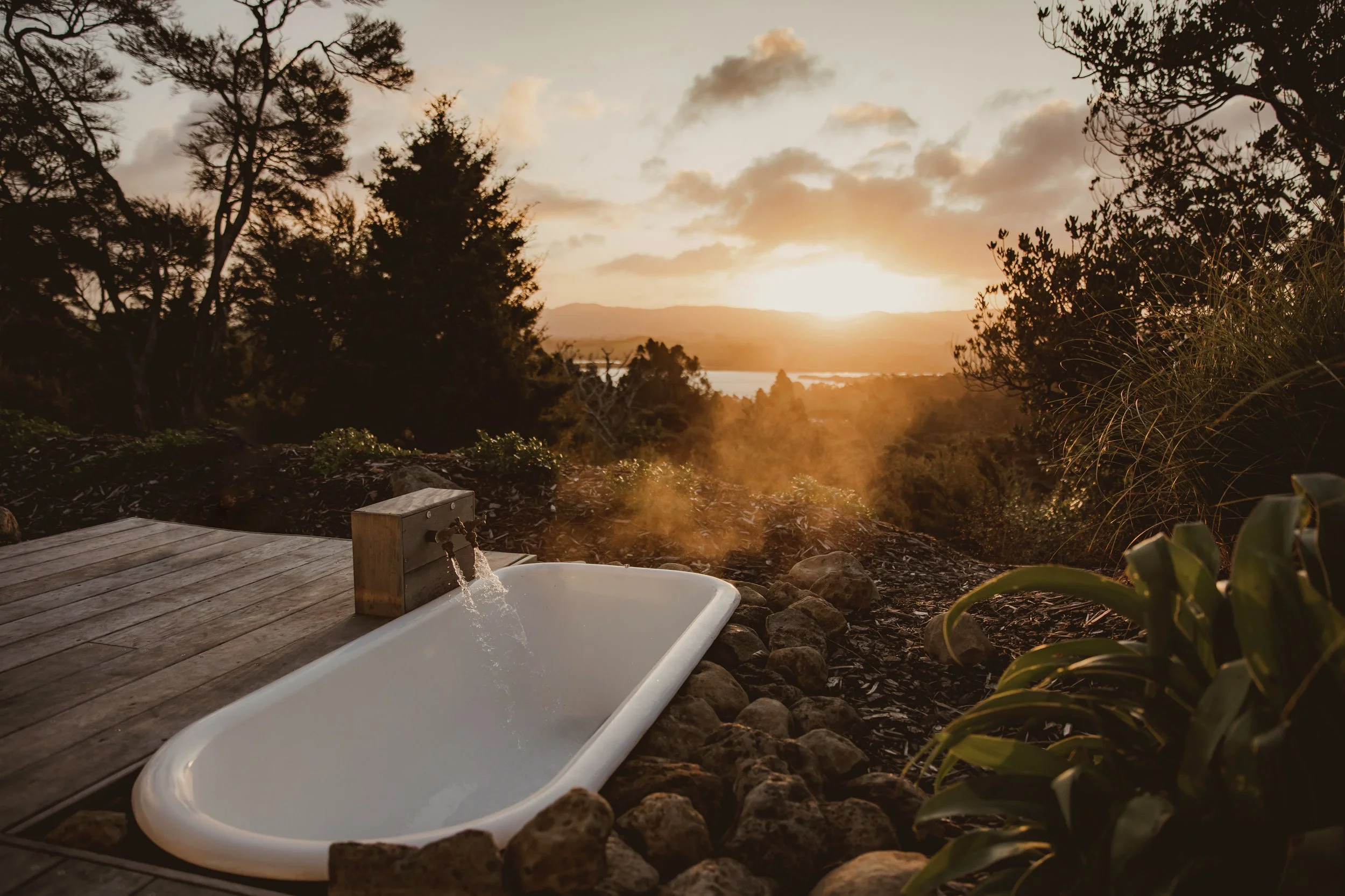 Outdoor bathtub with water flowing, surrounded by rocks and wooden decking, set in a natural landscape with a sunset view.