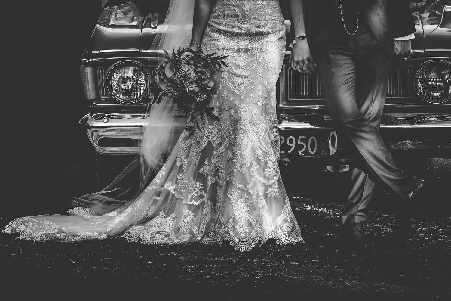 Bride and groom standing in front of a classic car, bride in a lace wedding dress holding a bouquet, groom in a suit, in a black and white photograph.