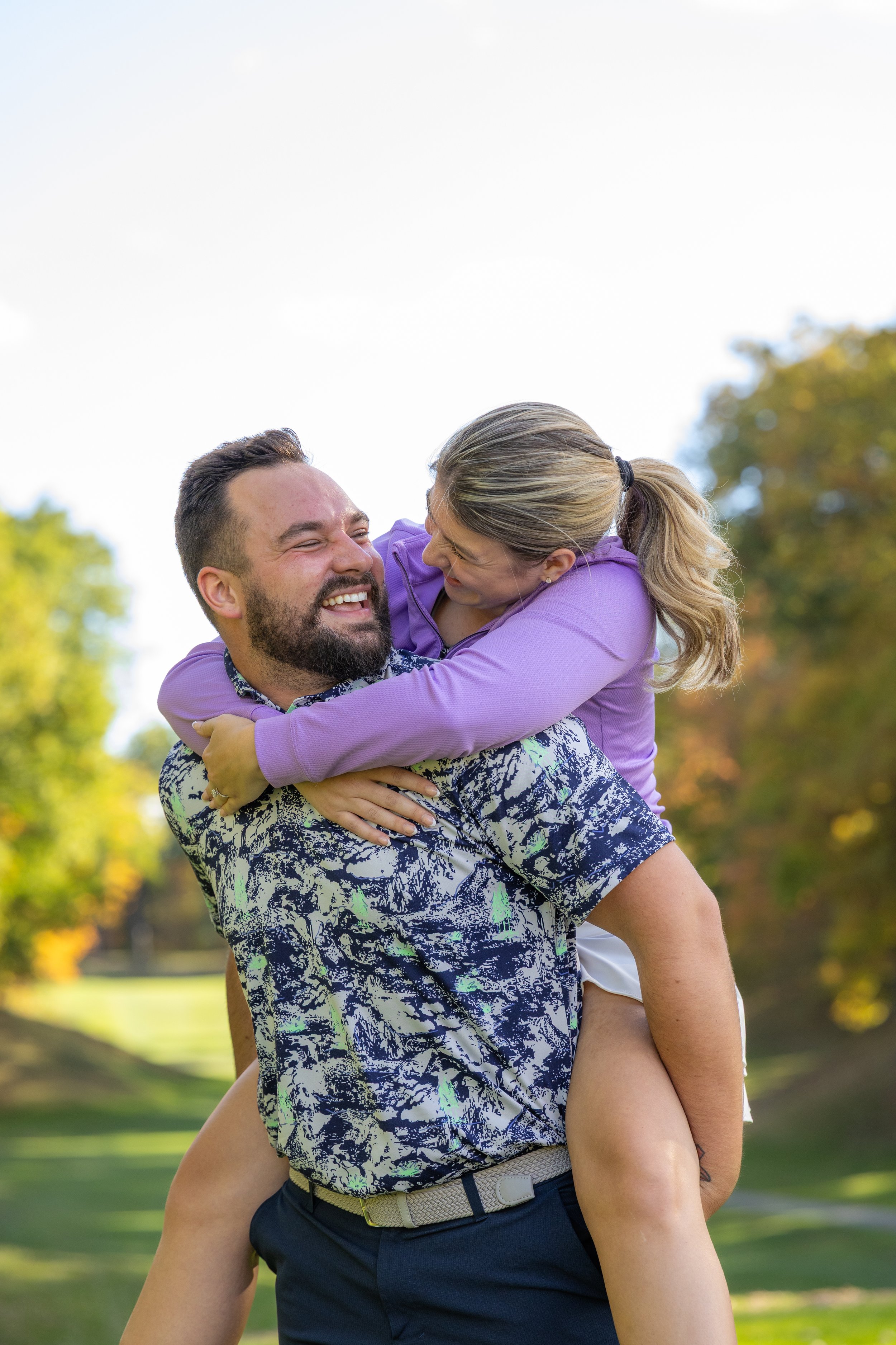 A man is carrying a woman on his back outdoors in a park, both smiling and enjoying each other's company with trees and blue sky in the background.
