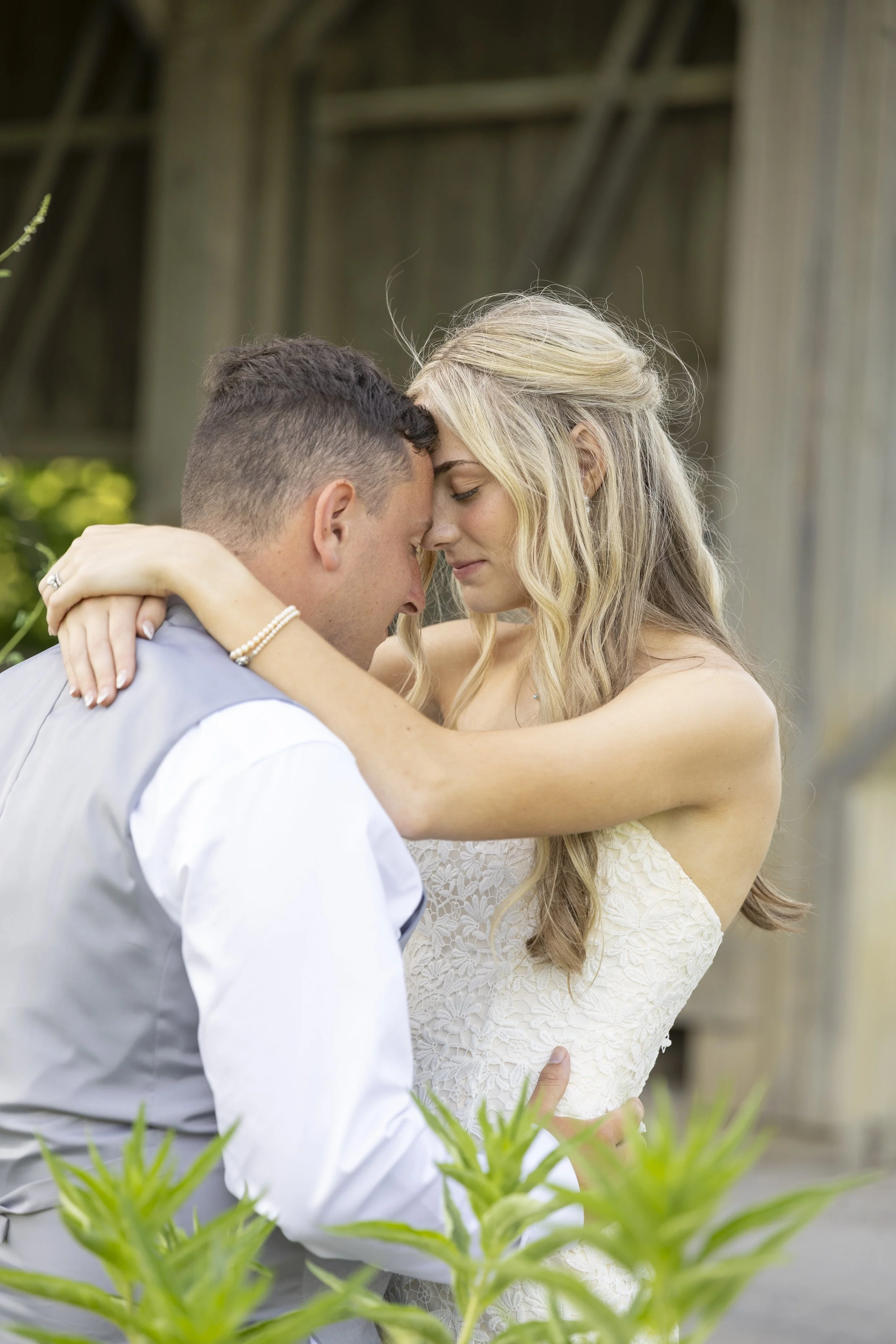 A couple shares an intimate moment with their foreheads touching outdoors, surrounded by greenery and a rustic wooden structure.