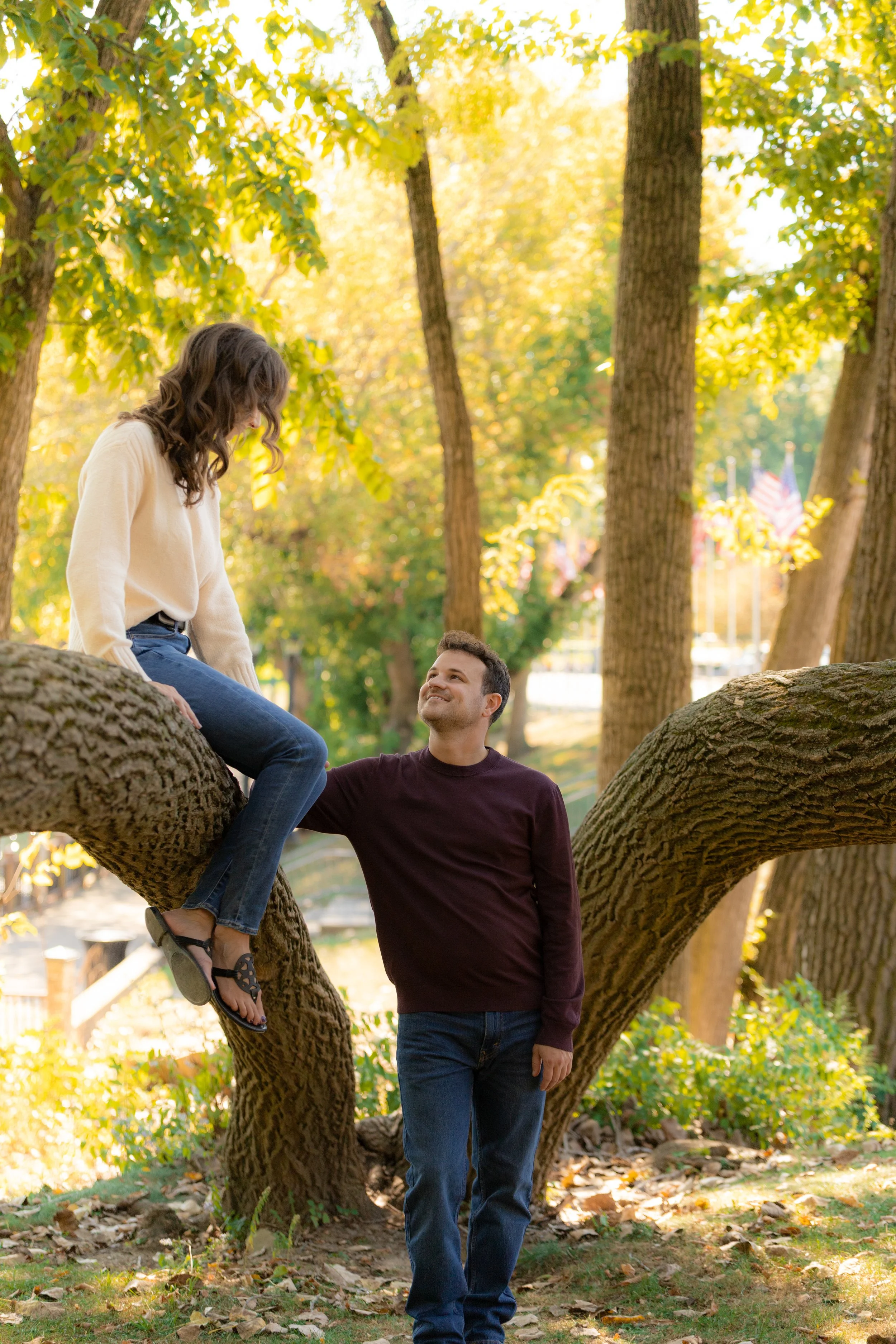 A man is standing holding a woman sitting on a tree branch in a park during autumn.
