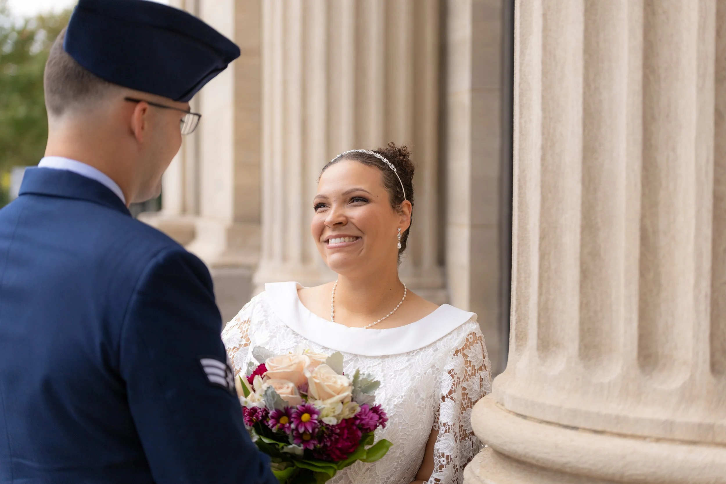 A bride in a white lace dress smiling at a man in a military uniform holding a bouquet of pink and white flowers.