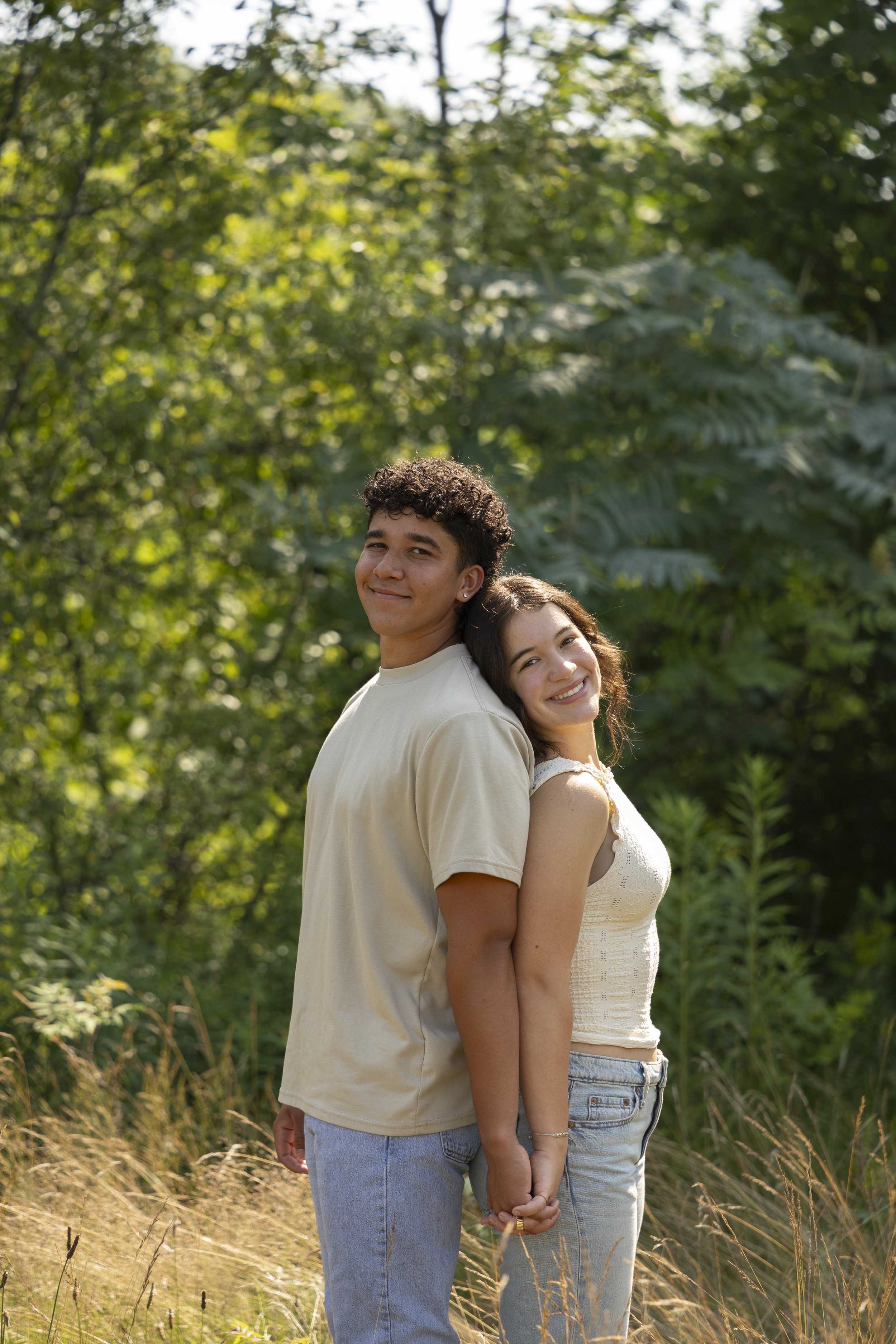 A young man and woman standing back-to-back outdoors holding hands, smiling, with lush green trees and tall grass in the background.
