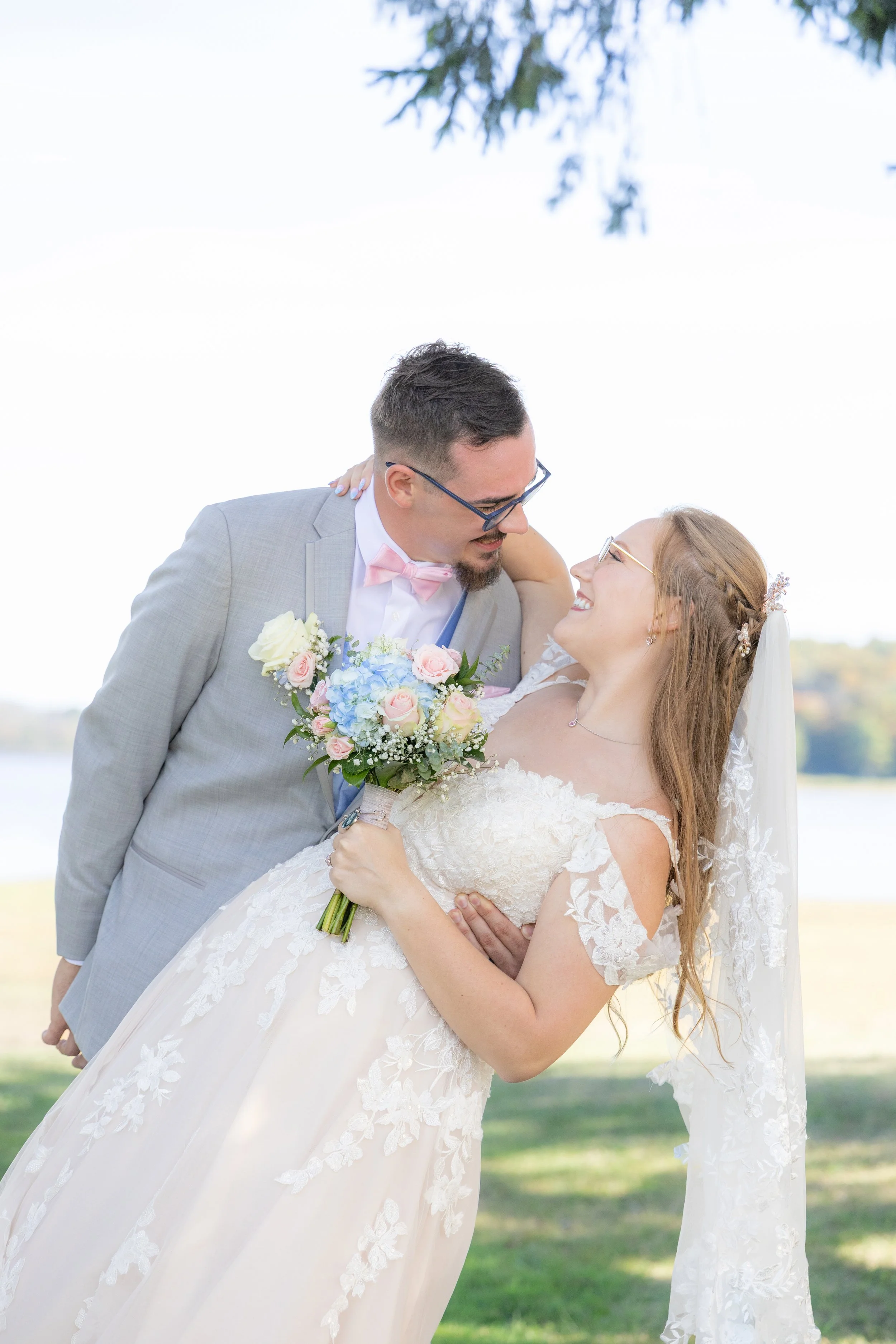 Bride and groom smiling and looking at each other outdoors, with the groom in a light gray suit and pink bow tie, and the bride in a lace wedding dress holding a bouquet of pastel roses and blue flowers.