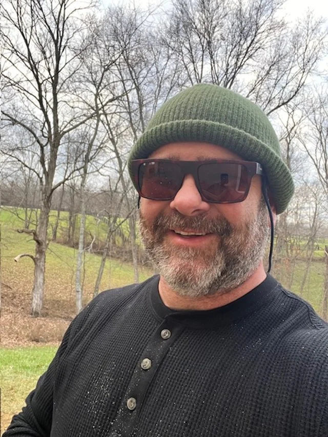 A man with a beard wearing sunglasses, a green beanie, and a black long-sleeve shirt smiling outdoors in a park with trees and grass in the background.