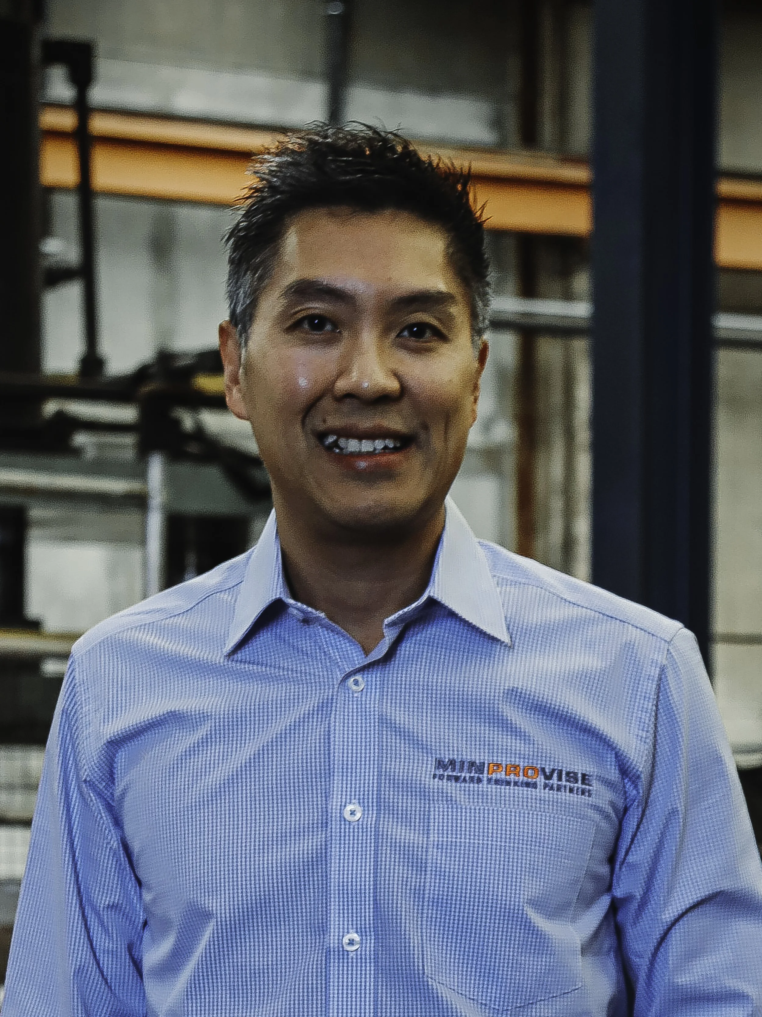 A middle-aged man with gray hair wearing a blue, collared, button-up shirt with a logo reading 'MINPROVISE' on the chest, standing indoors with a blurred background.