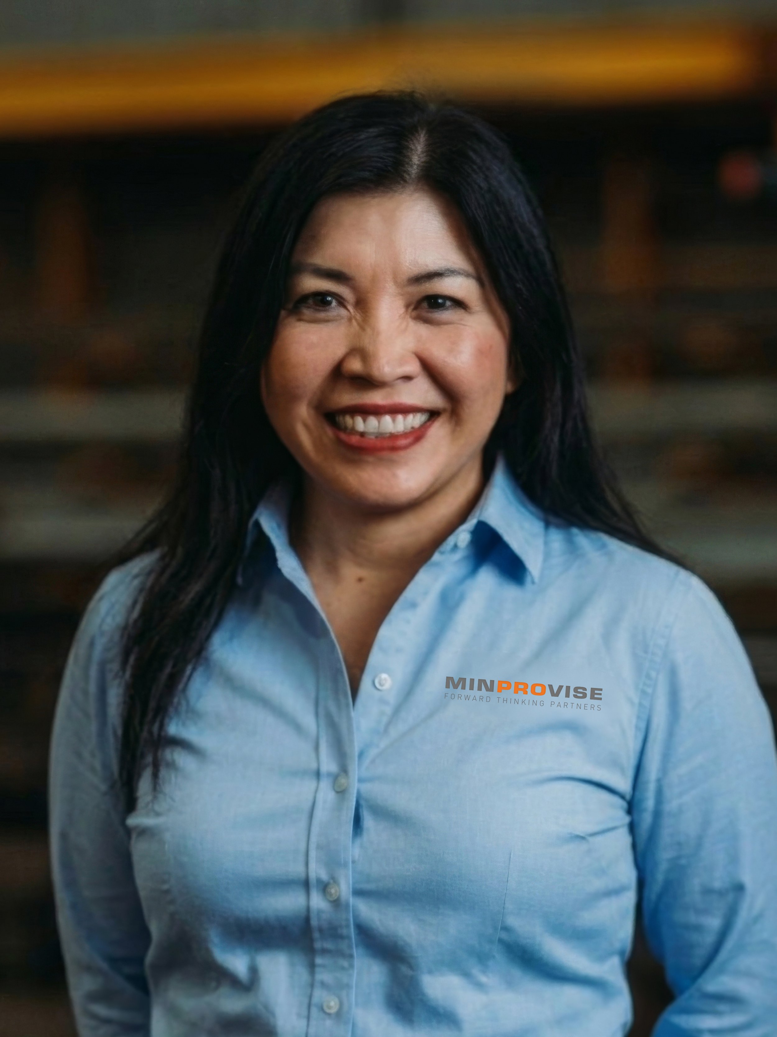 A woman smiling, wearing a dark shirt, standing in front of a blurred industrial or warehouse background.