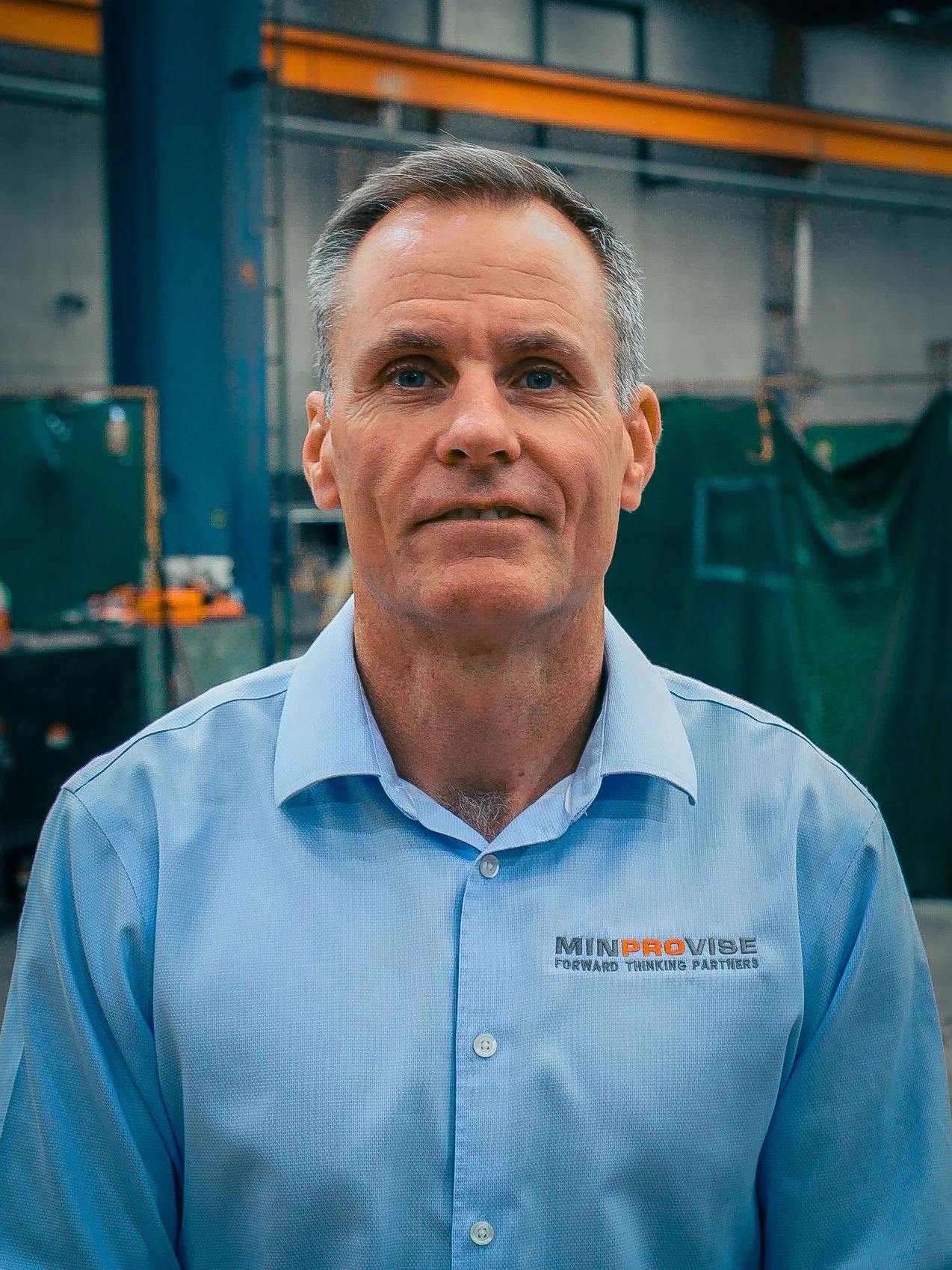 Man with short gray hair wearing a light blue dress shirt with 'MINPROVISE' logo, standing in an industrial or warehouse setting.