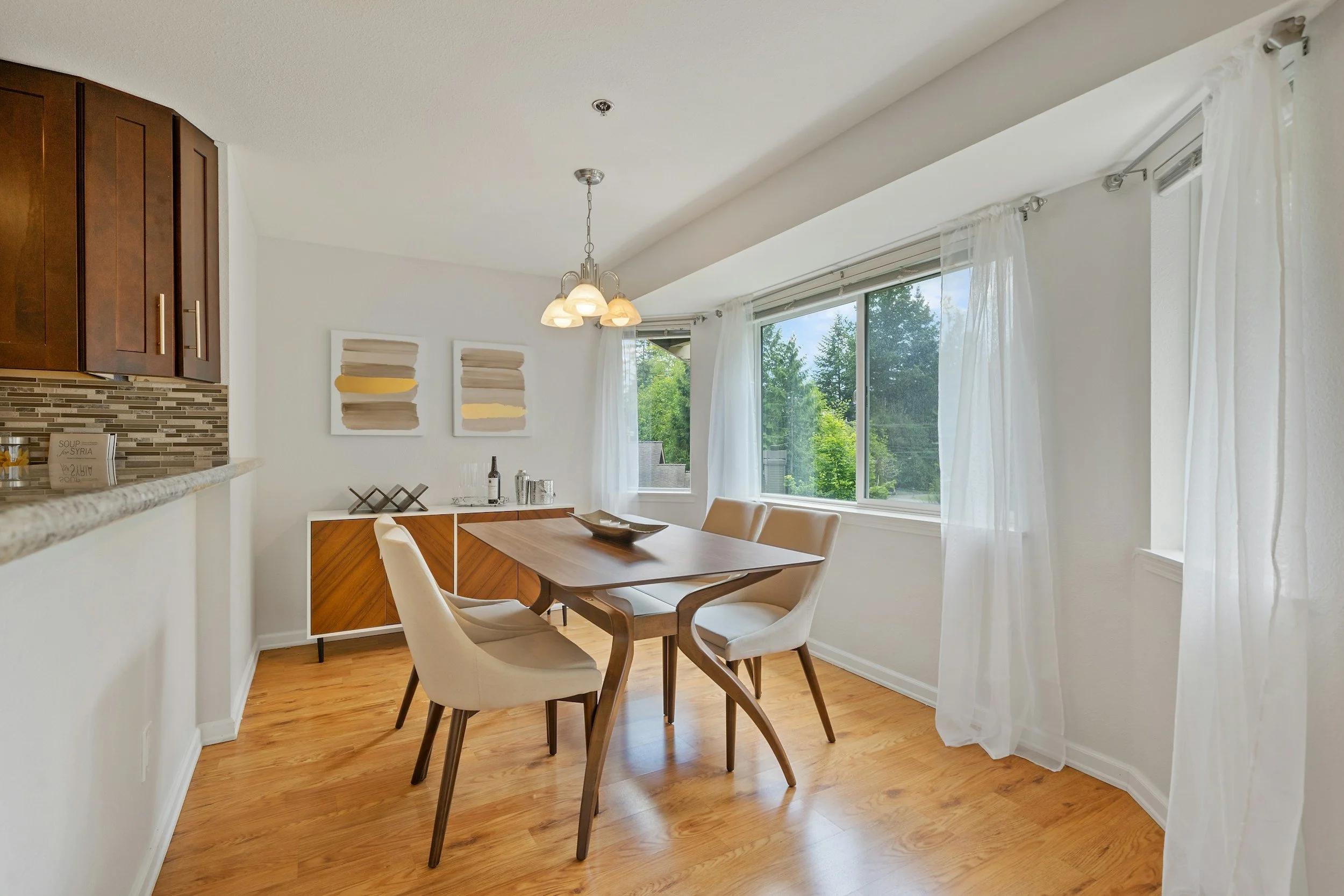 Dining room with a wooden table and four beige upholstered chairs, large windows with white curtains, a sideboard with art pieces and wine bottles, hardwood floors, and a hanging light fixture.