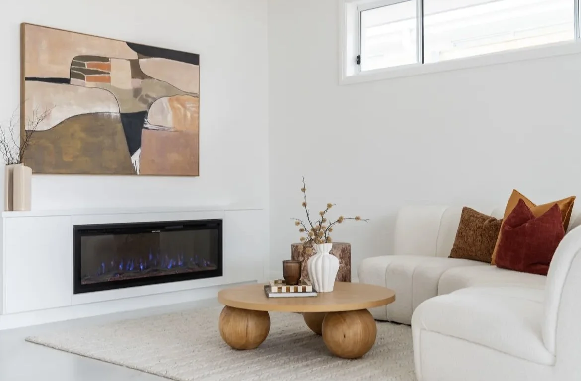 Modern living room with a white sofa, colorful pillows, wooden coffee table with decorative vases, abstract wall art, built-in fireplace with blue flames, neutral-colored rug, and a small window