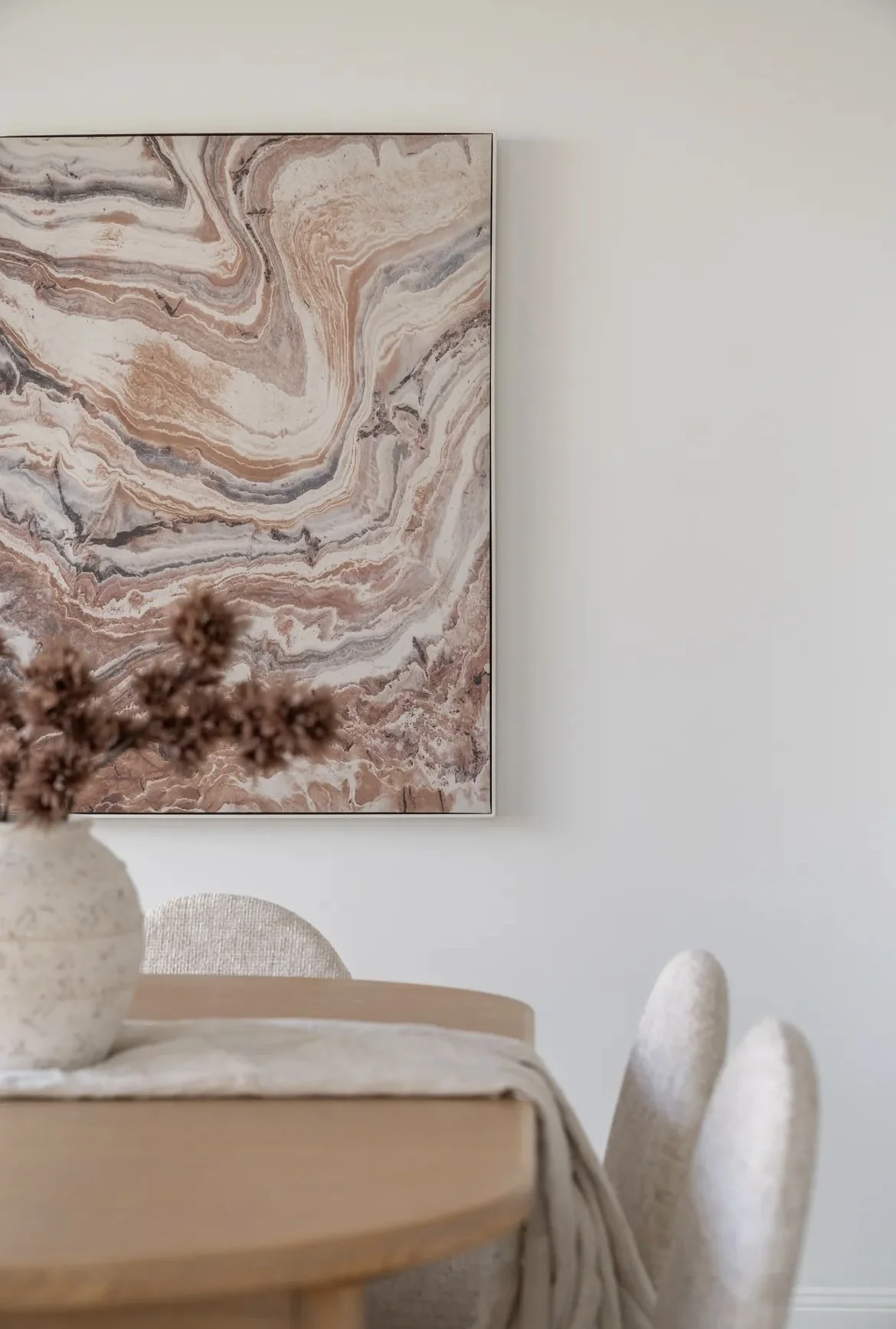 A modern dining room with a round wooden table, two upholstered chairs, a marble-patterned painting on the wall, and a vase with dried flowers on the table.