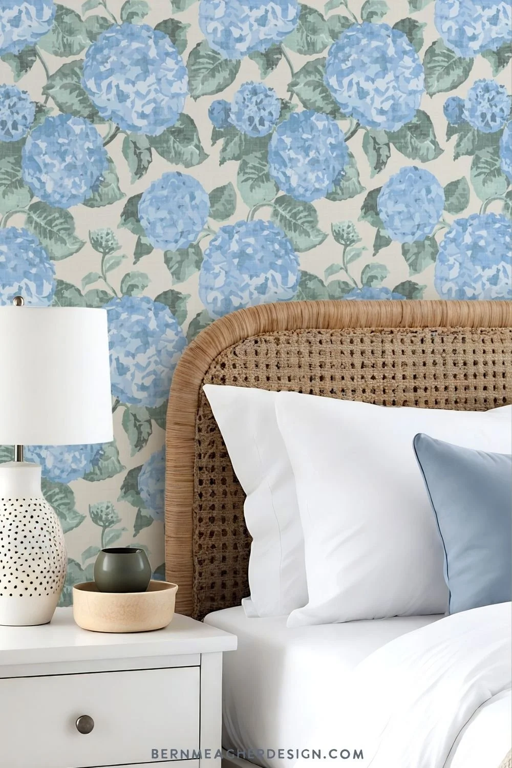 Coastal grandmother style french blue bedroom with French blue hydrangea peel and stick wallpaper, rattan cane headboard, white bedding, blue accent pillow, and white nightstand with ceramic lamp and decor.