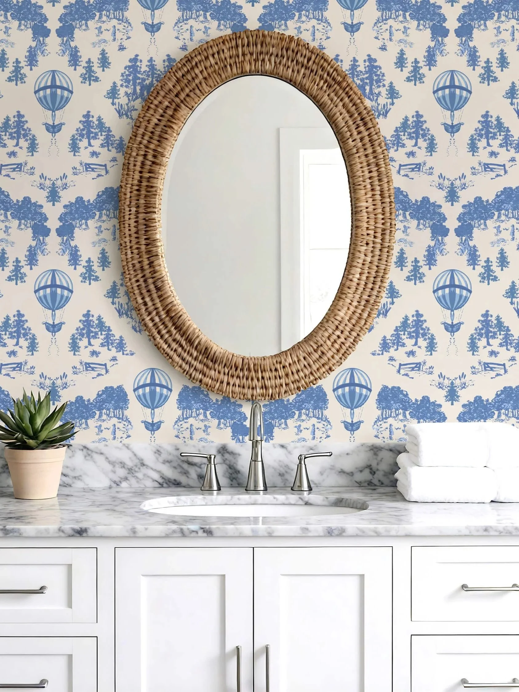 A bathroom with a light blue floral and hot air balloons floating over countryside patterned wallpaper, behind a rustic rattan mirror and white marble top vanity with plant