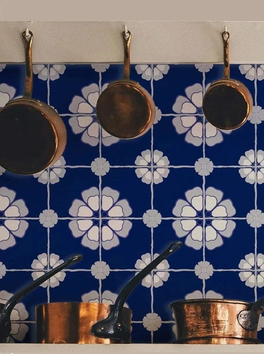 Copper pots hanging on a wall above a kitchen counter with black handles, set against a blue and white floral patterned faux tile wallpaper backsplash.