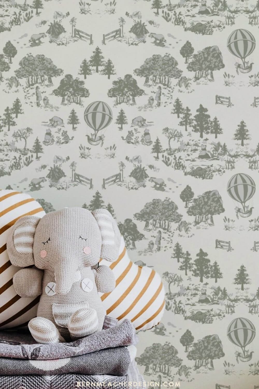 Peel and stick winnie the pooh countryside nursery wallpaper with olive green woodland toile scenes and hot air balloons on a cream background, styled behind a stack of folded blankets and a knitted gray elephant toy with a striped pillow.