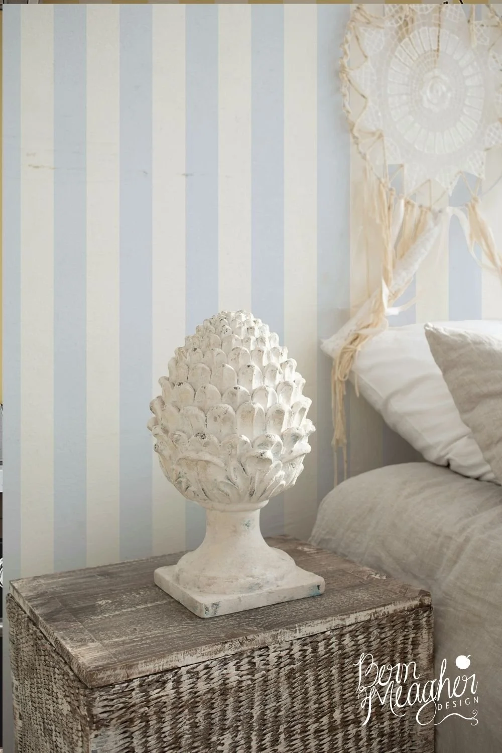 Pale blue and cream vertical stripe wallpaper in a coastal bedroom setting featuring a wicker side table topped with a white decorative sculptural object and neutral-toned bedding with a crochet wall hanging in the background.