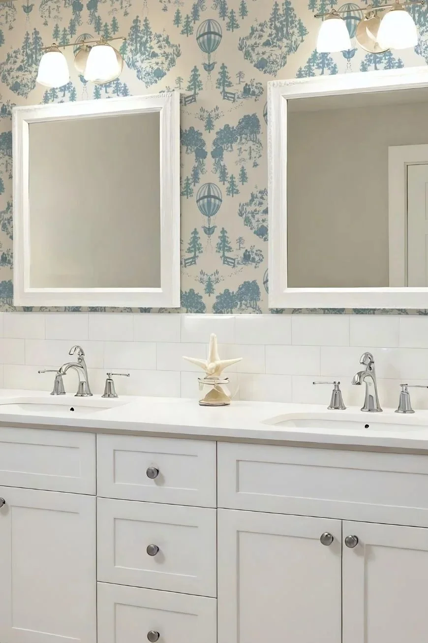 Blue and creamy white traditional toile vinyl wallpaper with hot air balloons over scenic countryside images behind white sconces, a white bathroom cabinet  with marble top and silver tapware