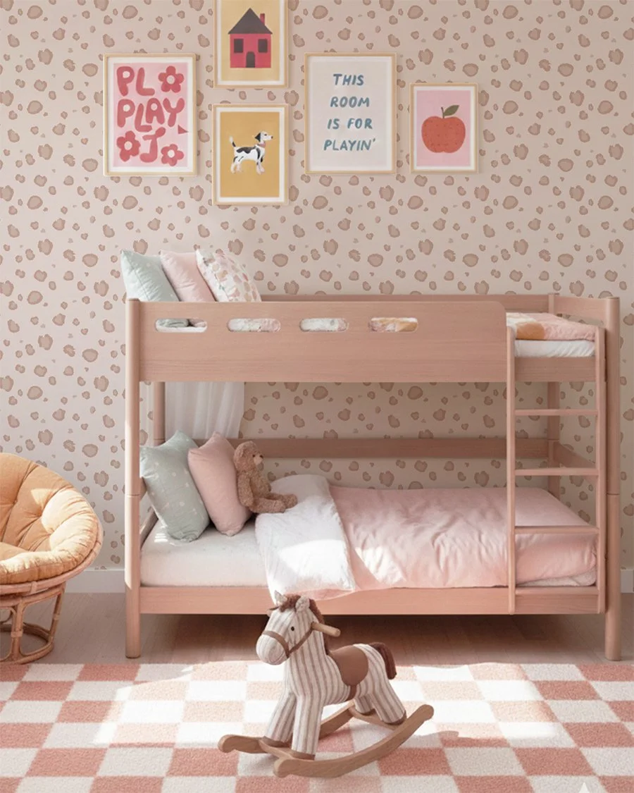 Soft pink whimsical wallpaper with mellow animal spots pattern in a girls bedroom nursery featuring a wooden bunk bed, pastel pillows, framed playful art prints, and a striped rocking horse toy on a checkered rug.