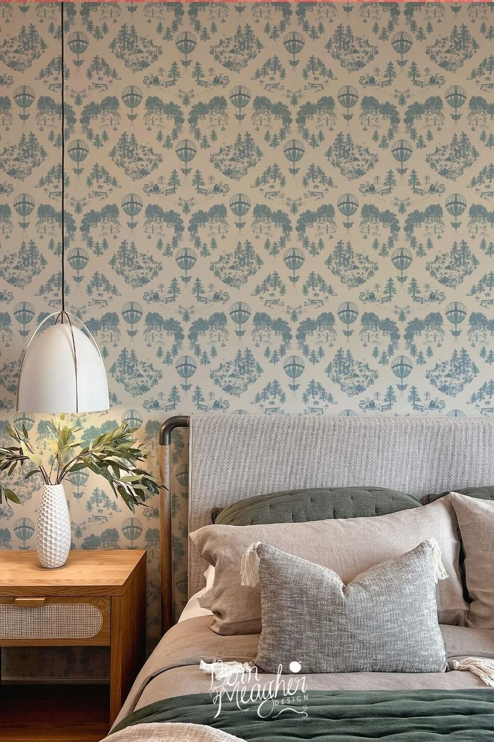 Blue and white traditional toile wallpaper with balloons and scenic park images behind a cozy bedroom setup with a grey upholstered headboard, neutral-toned bedding, and a wooden bedside table with a vase of greenery and a hanging white dome lamp.