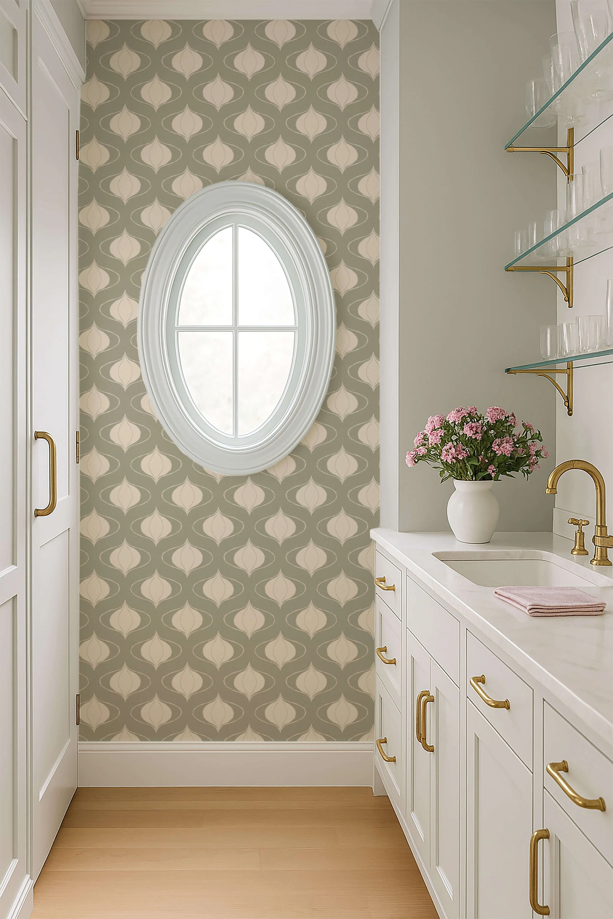 Peel and stick wallpaper in a soft sage green and cream palette featuring a repeating, symmetrical botanical-inspired pattern in Arts & Crafts style. in a bright kitchen with white cabinetry, brass hardware, glass shelves, and a white oval window.