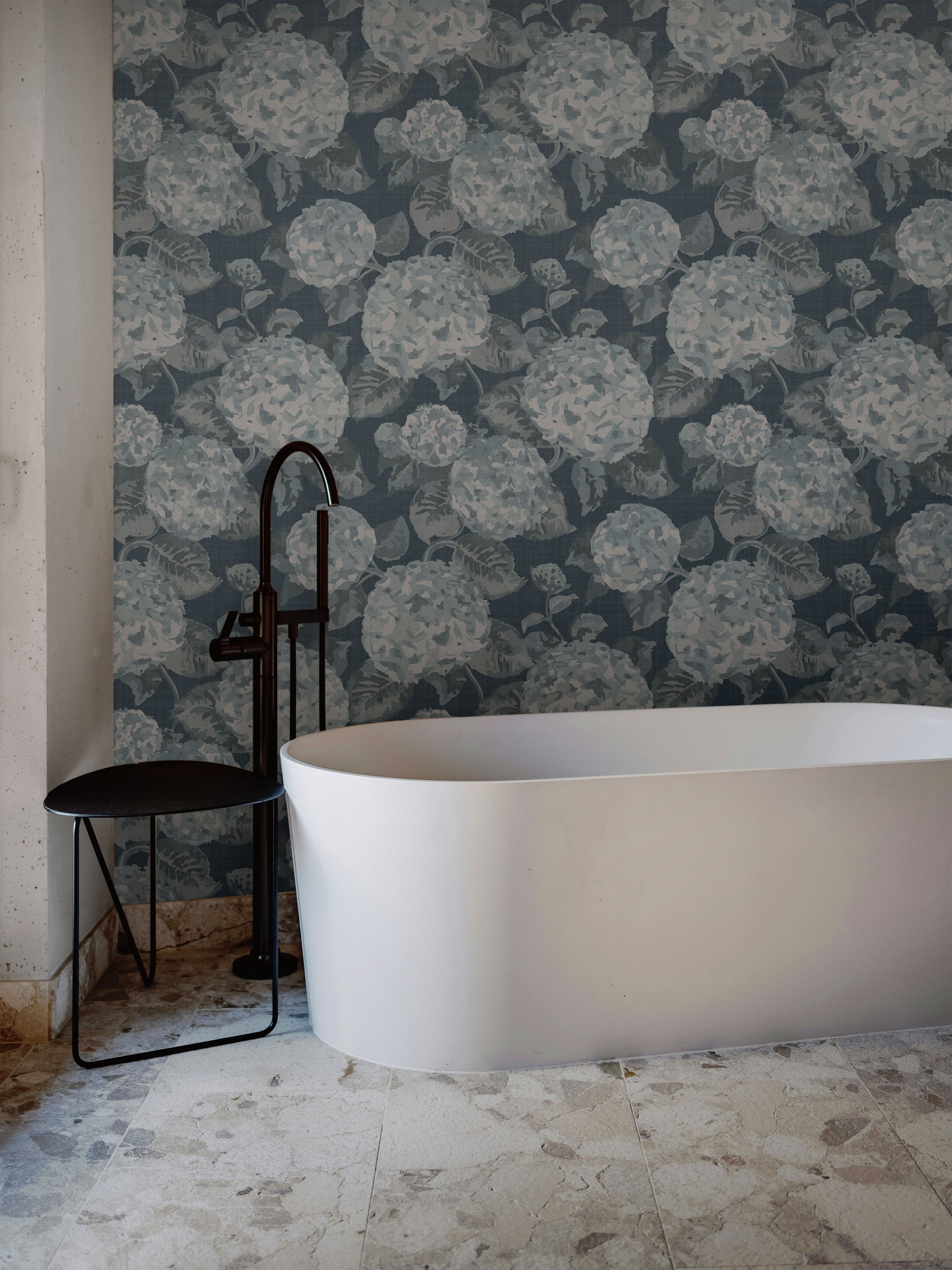 Moody blue hydrangea floral vinyl wallpaper on a bathroom feature wall behind a white freestanding tub, with stone tile floor and black faucet and stool.