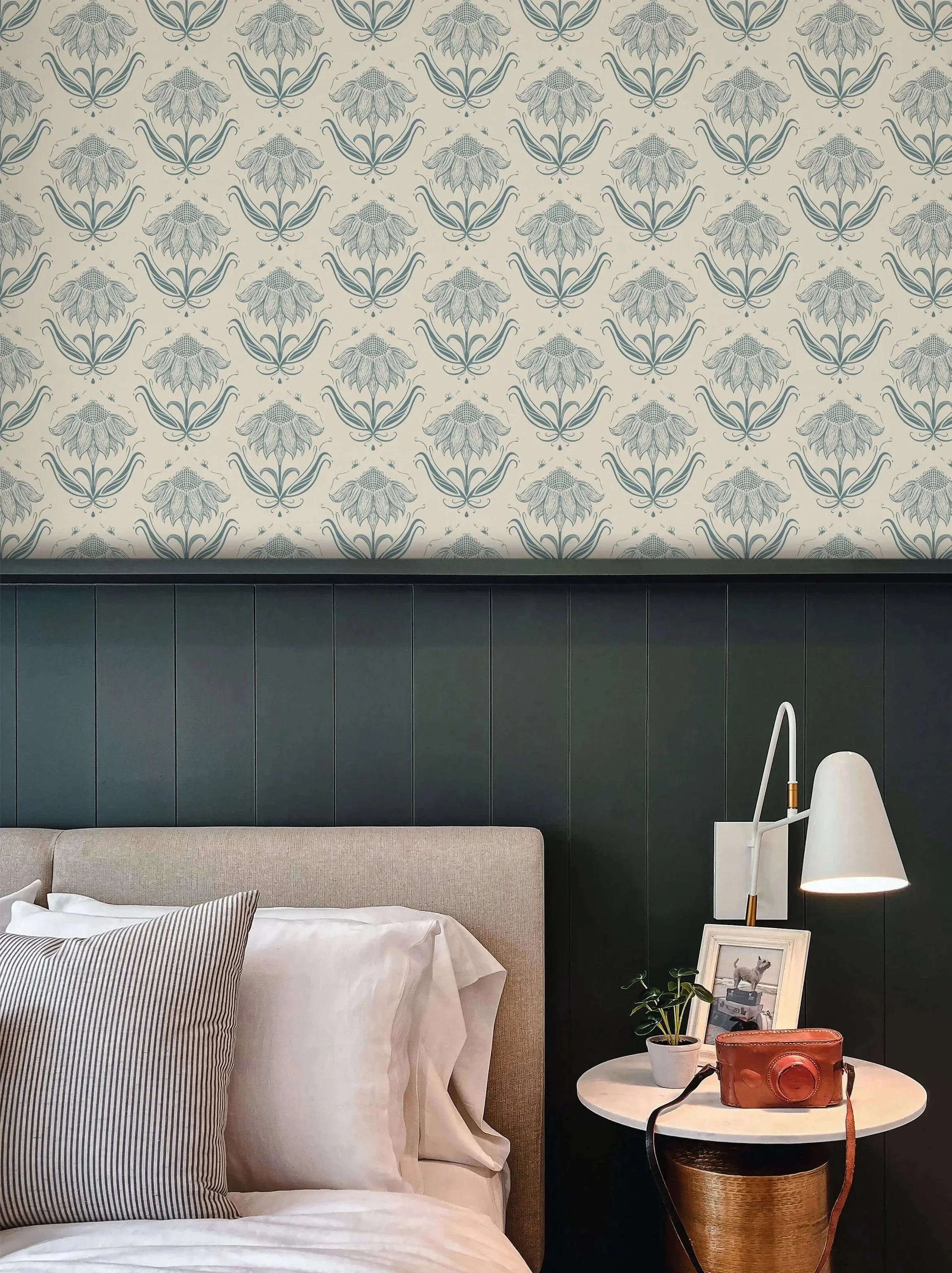 Interior bedroom scene featuring sunflower with bees wallpaper, with a bed, pillow, bedside table, framed photo, plant, and lamp.