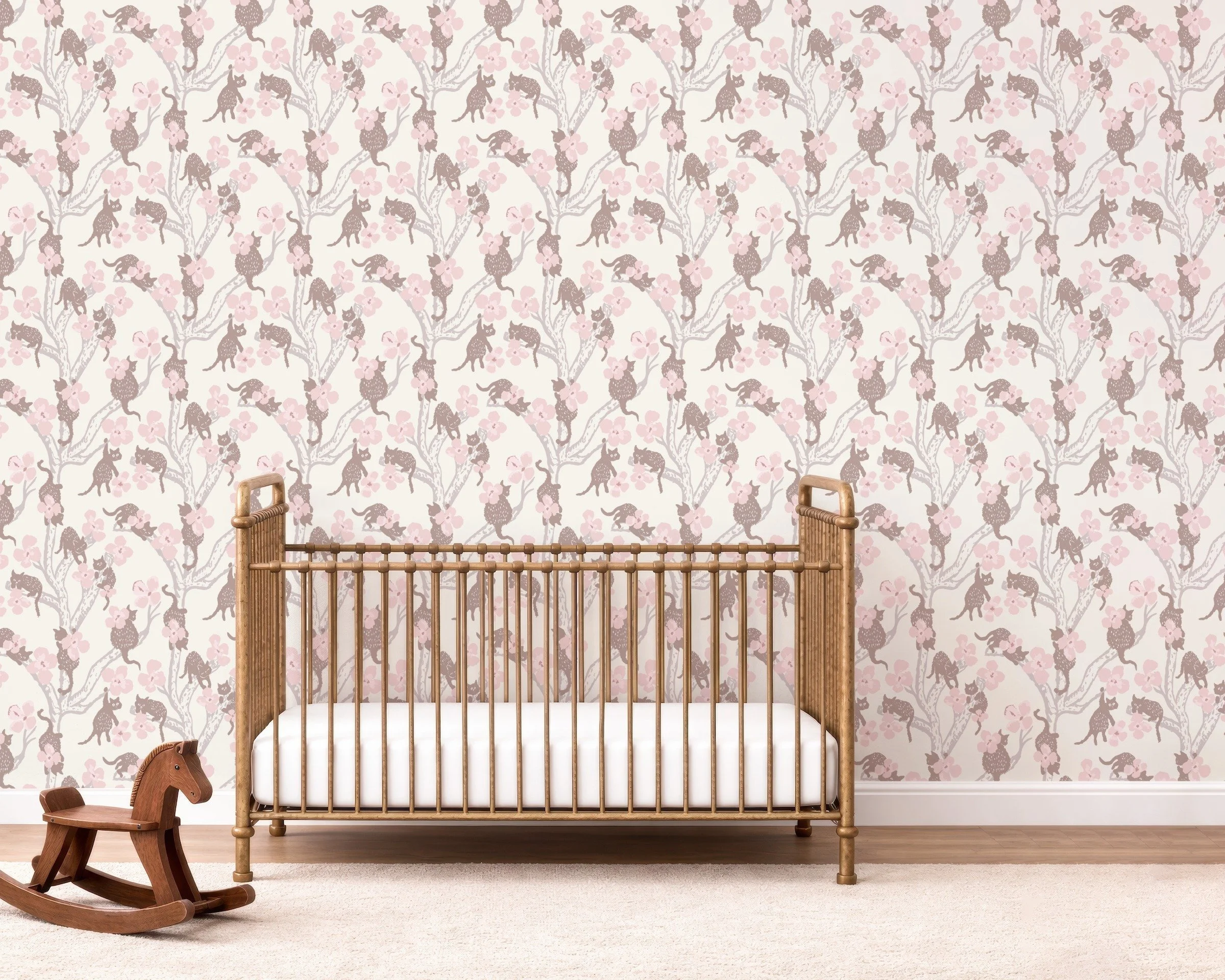 Blush pink floral and vintage pink cat peel and stick nursery wallpaper behind a natural wood crib and wooden rocking horse on a light rug in a soft, neutral baby room.