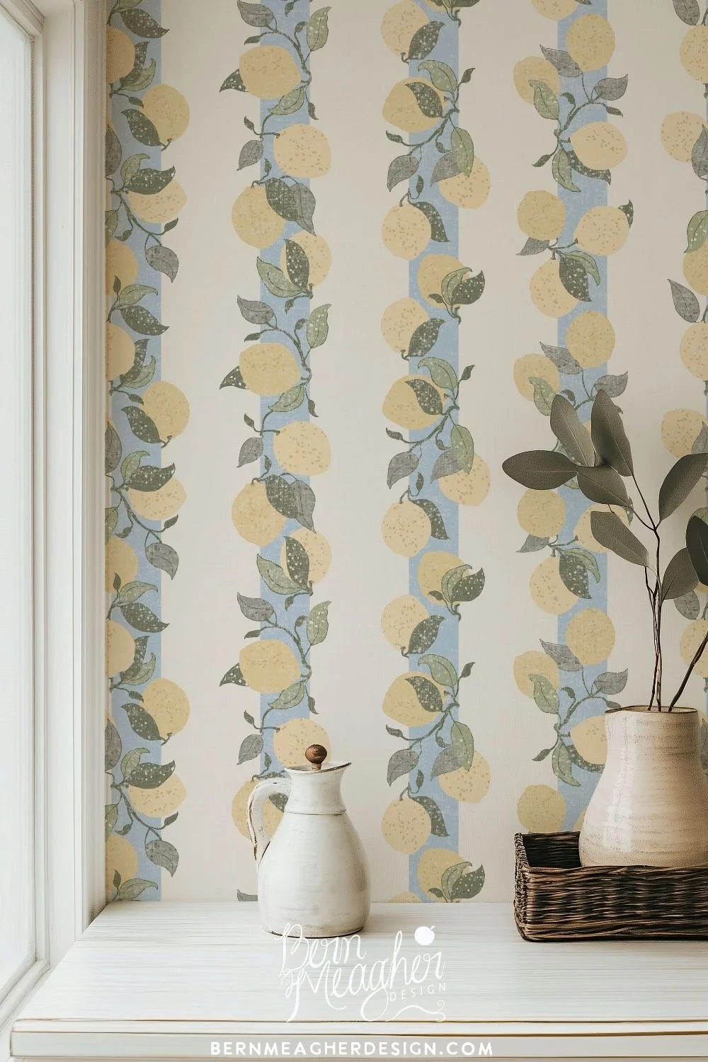 Farmhouse-style wallpaper in warm muted butter yellow and sage greens with textured blue vertical stripes featuring citrus lemons and leafy branches, shown in a bright kitchen nook with white cabinetry and ceramic vases on a white wooden countertop.