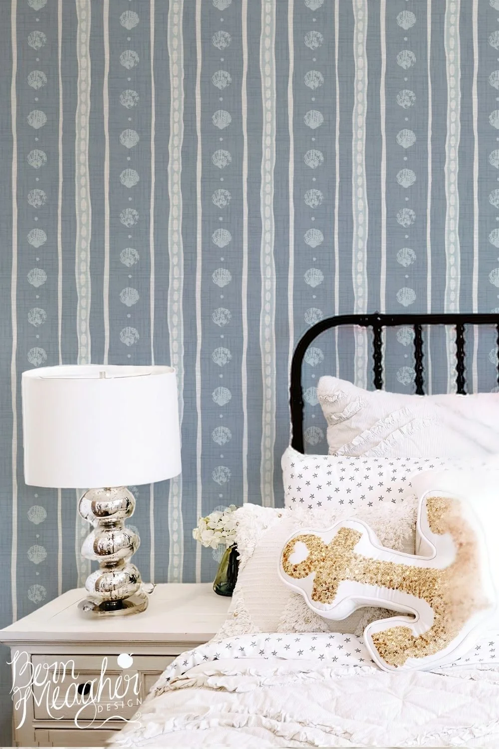 Soft blue and white peel and stick wallpaper with block print shells and organic white stripes behind a white bed with plush beige anchor cushio. A white lamp with silver stand sits beside bed on pale wood table